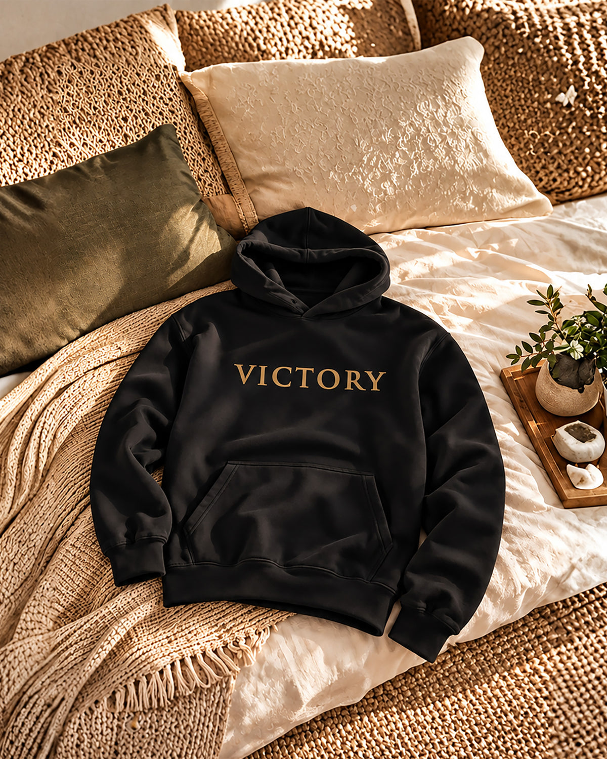 A black pullover hoodie with the word 'VICTORY' printed in gold across the chest, styled on a bed with beige bedding, woven pillows, and a small plant.