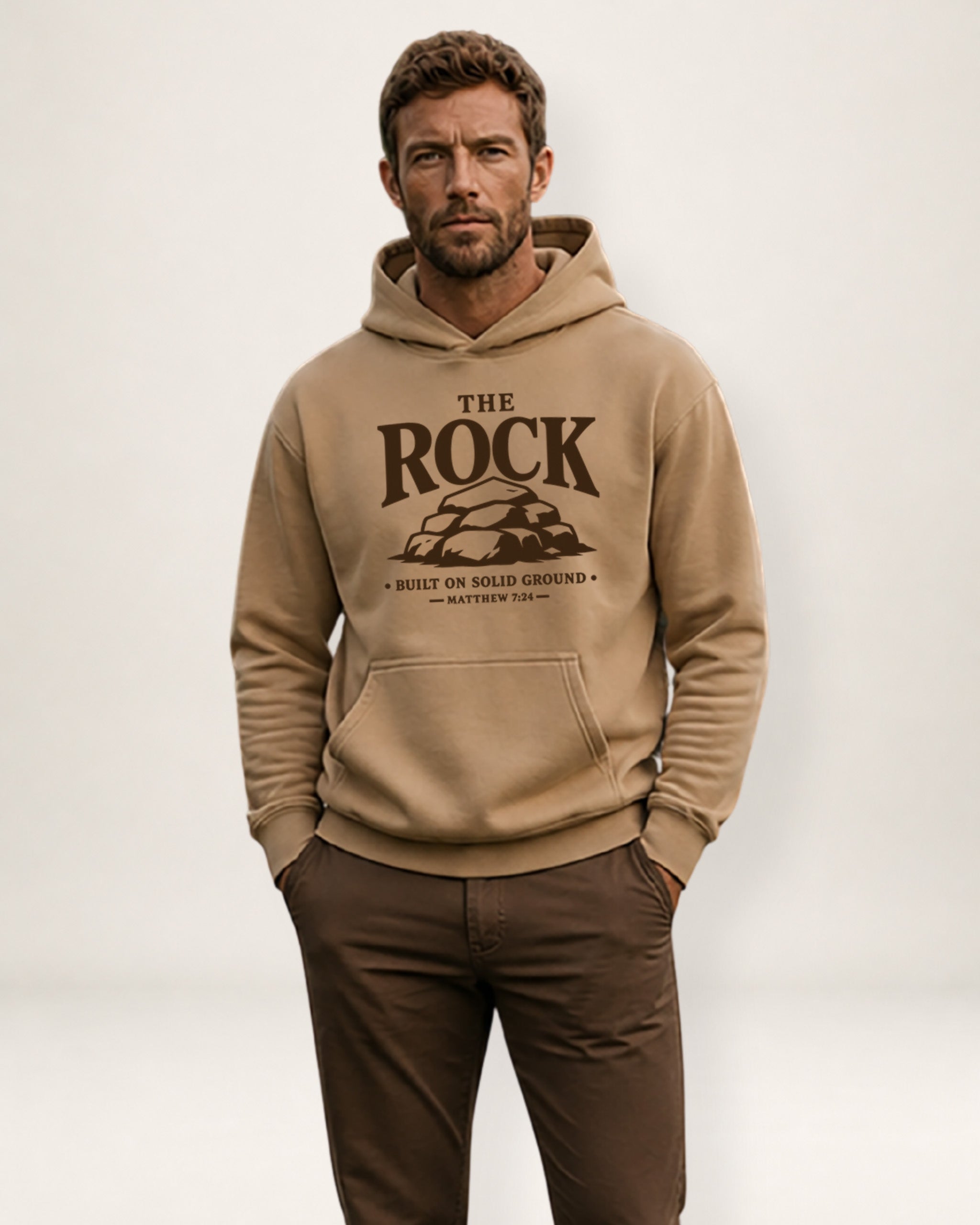 A man wearing a beige pullover hoodie with a dark brown graphic print that reads 'THE ROCK BUILT ON SOLID GROUND ANTHONY 2013' and matching brown pants, standing against a light gray studio background.
