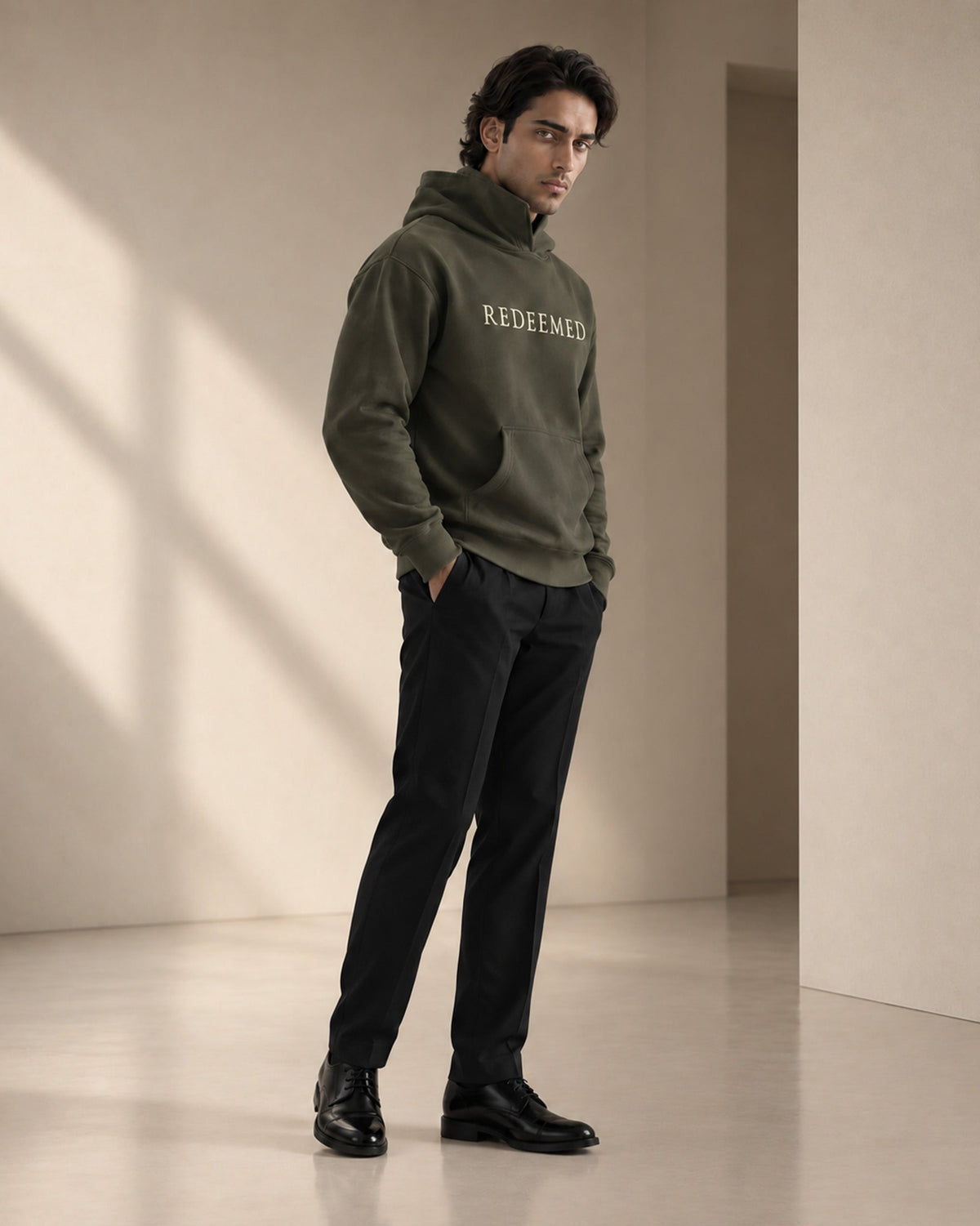 A man wearing an olive green pullover hoodie with 'REDEEMED' printed in white across the chest, paired with black trousers and black dress shoes, standing in a minimalist beige room with soft window shadows.