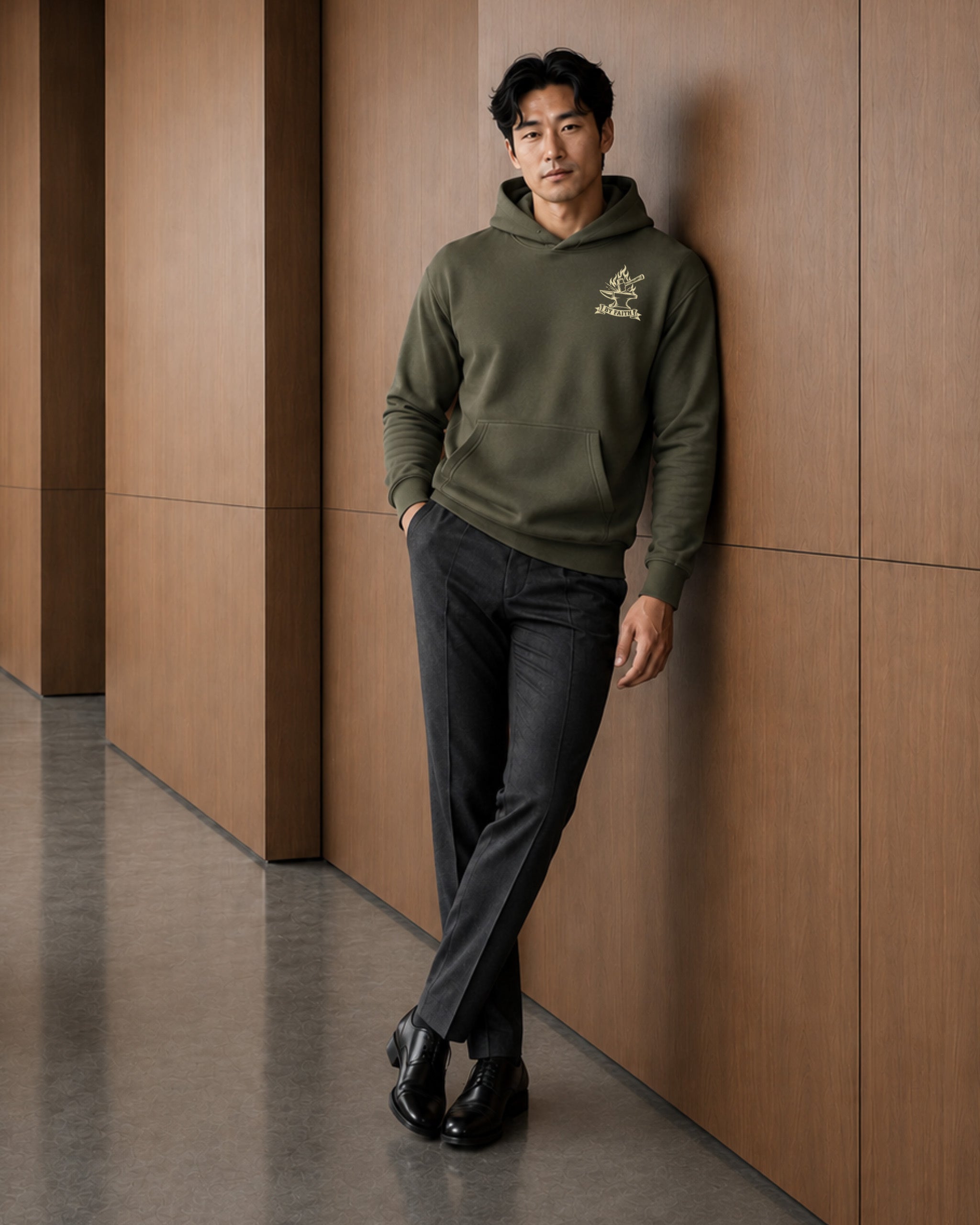 A man wearing an olive green hoodie with a small embroidered logo on the left chest, paired with dark gray trousers and black dress shoes, leaning against a modern wood-paneled wall.