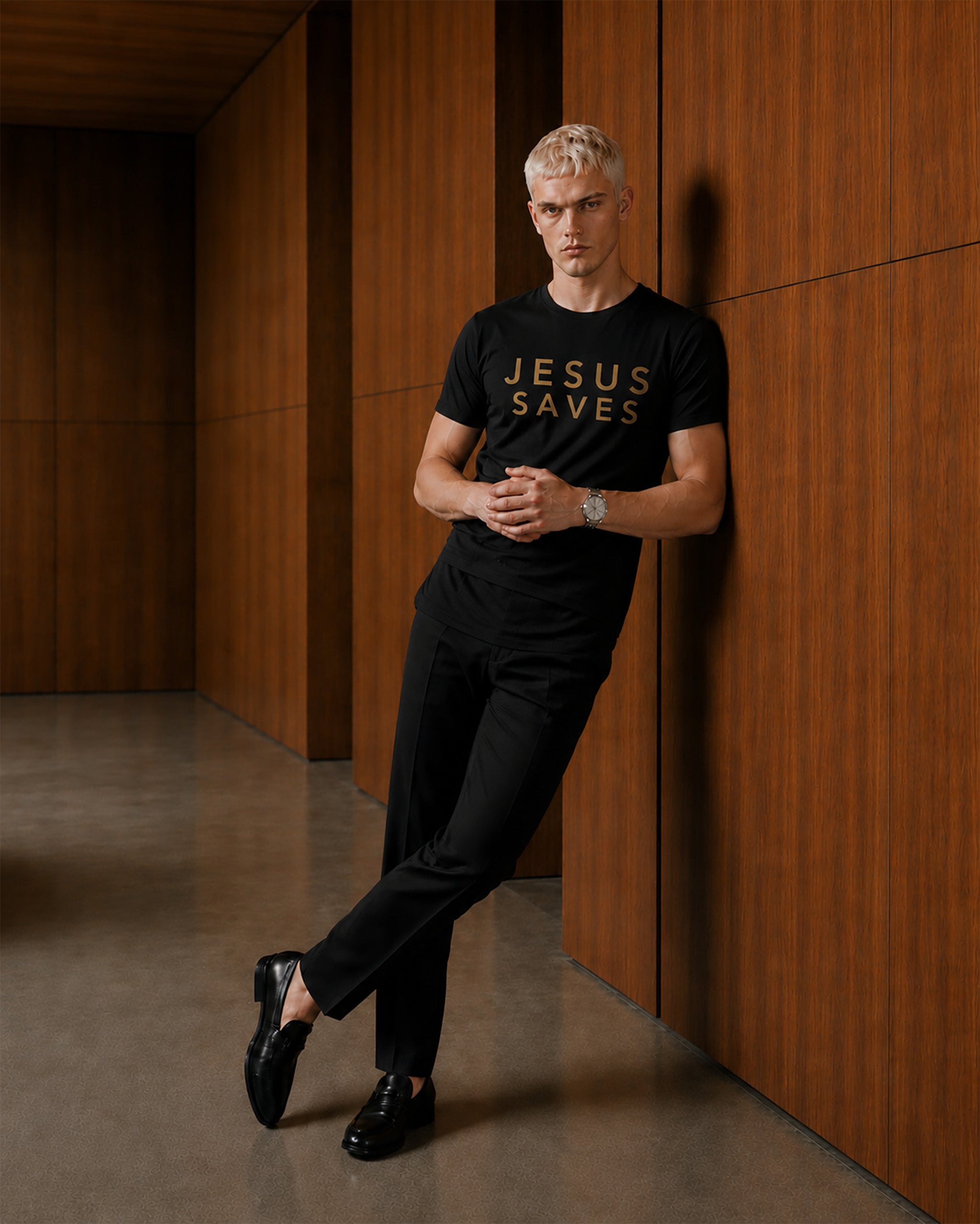 Man wearing a black t-shirt with 'JESUS SAVES' text, standing against a wooden wall.