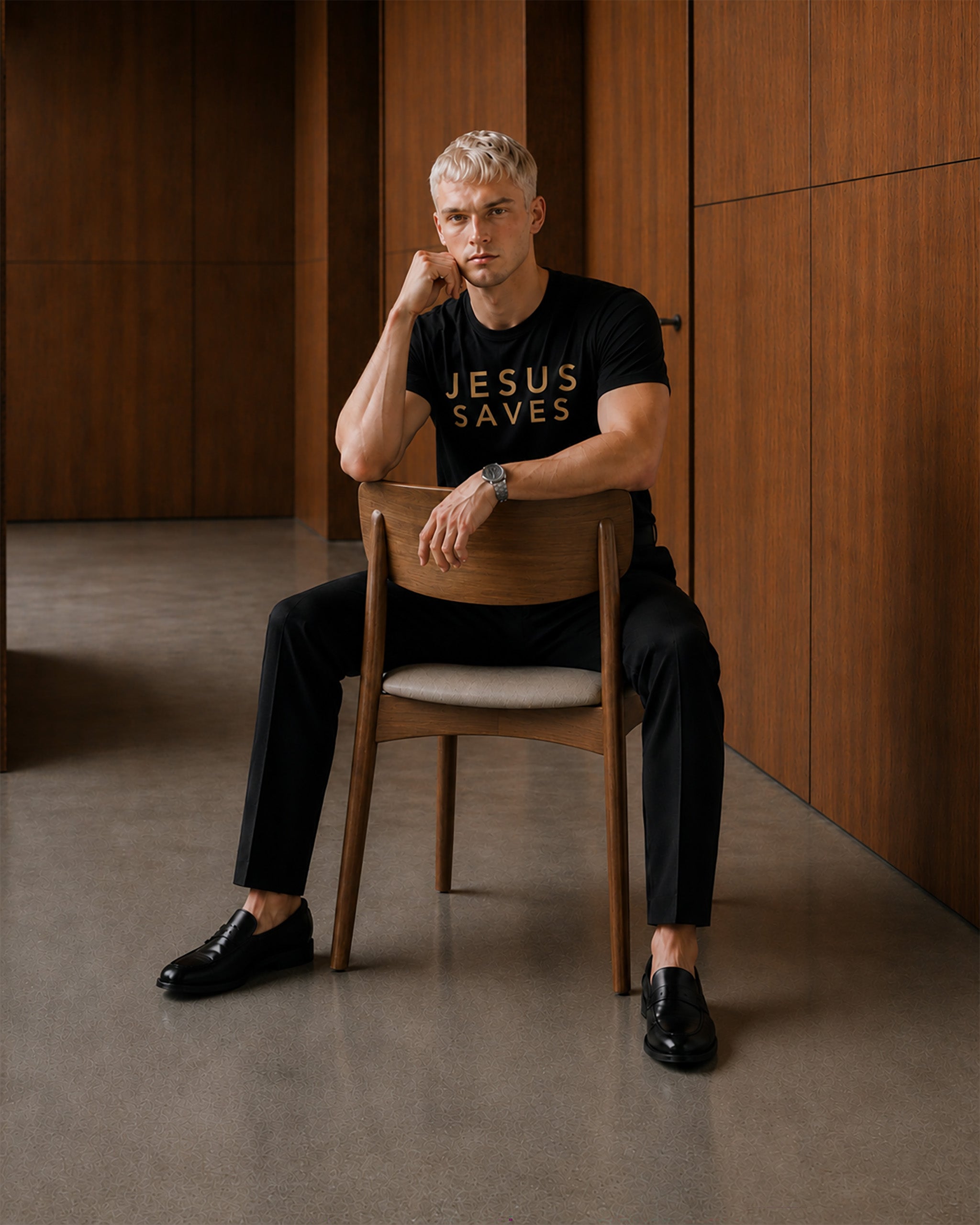 Man sitting on a chair wearing a black t-shirt with 'JESUS SAVES' text in a minimalistic room.