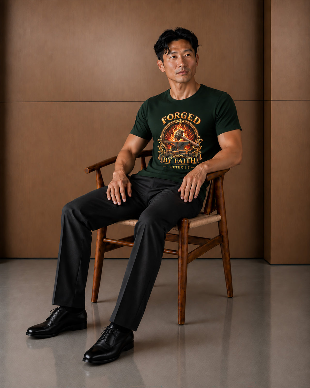 A man sits on a wooden chair wearing a dark green graphic t-shirt with 'FORGED BY FAITH' printed on the front, paired with black trousers and black dress shoes, set against a warm brown paneled wall.