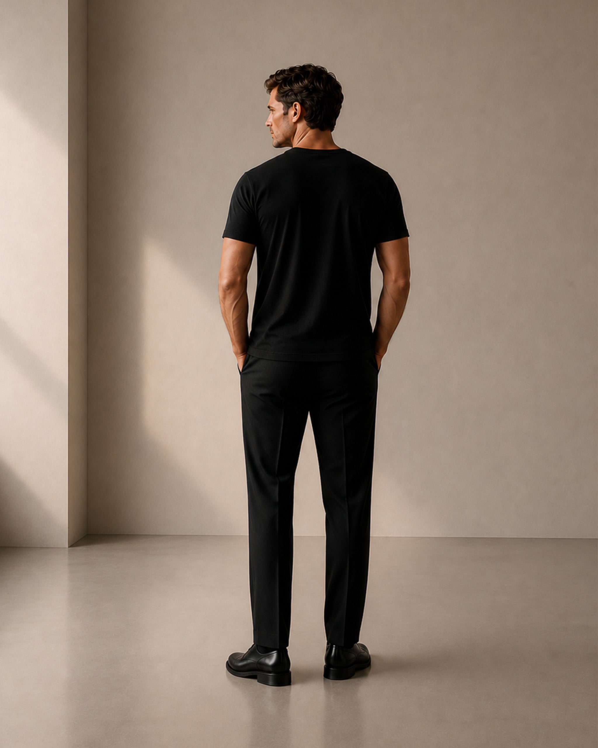 A man stands with his back to the camera wearing a black short-sleeve crewneck t-shirt and black trousers in a minimalist studio setting with warm beige walls and floor.
