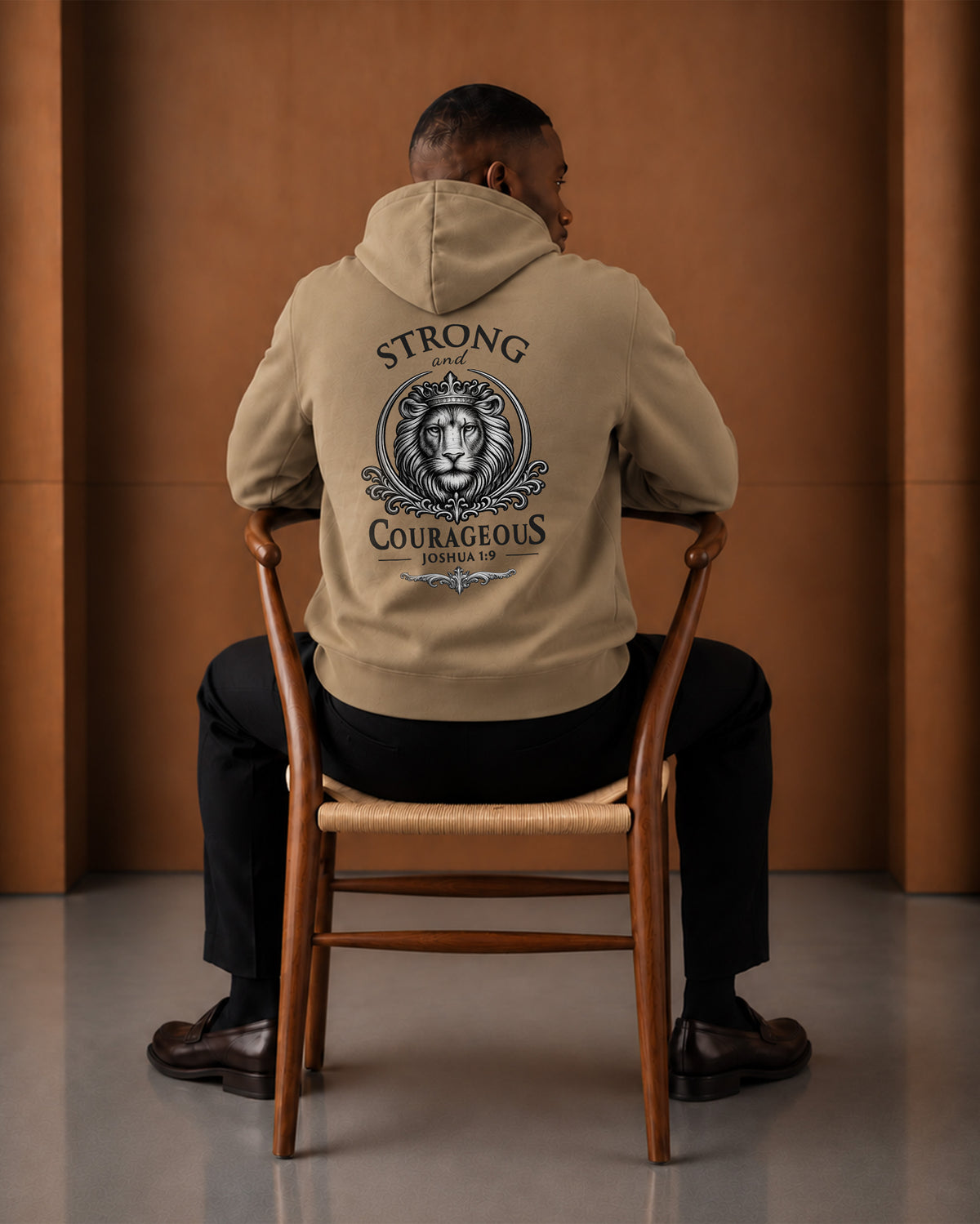 Seated rear-view styling of the Strong & Courageous sand hoodie, showing the hood and back artwork.