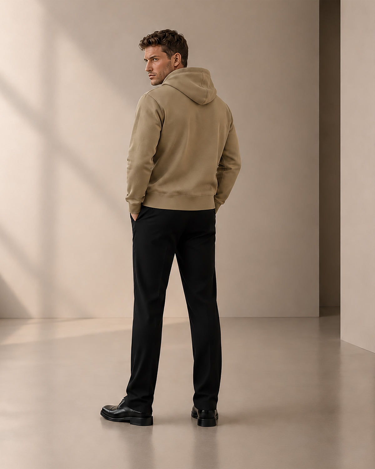 Rear view of a man wearing the sand SEALED hoodie and black trousers, showing the clean back and drawcord-free hood in a warm minimalist studio.