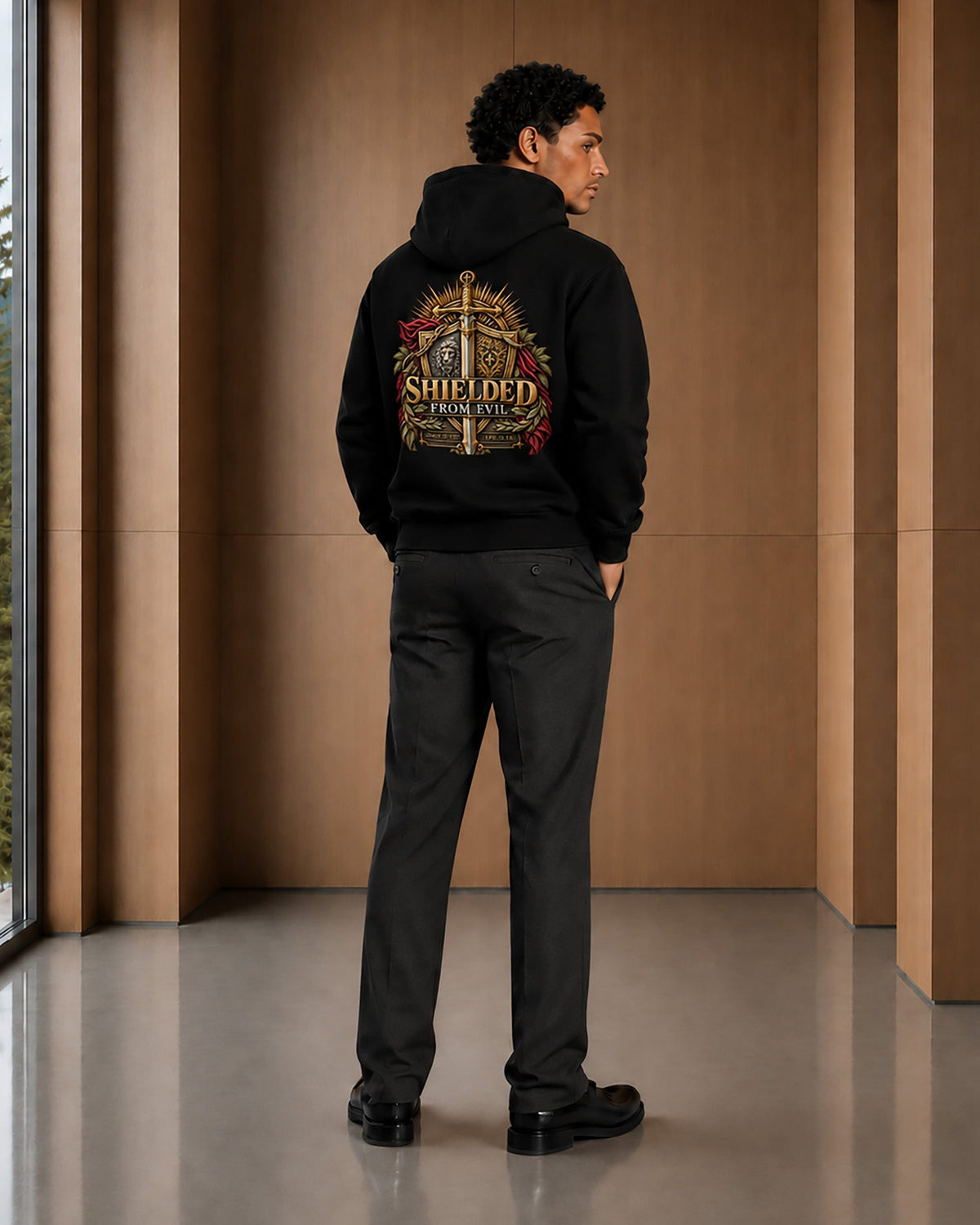 A man stands in a modern indoor setting with wood-paneled walls, viewed from behind. He is wearing a black hoodie with a large, detailed graphic on the back featuring a sword, shield, and the text 'SHIELD OF NEW ENGLAND,' along with dark gray pants and black shoes.