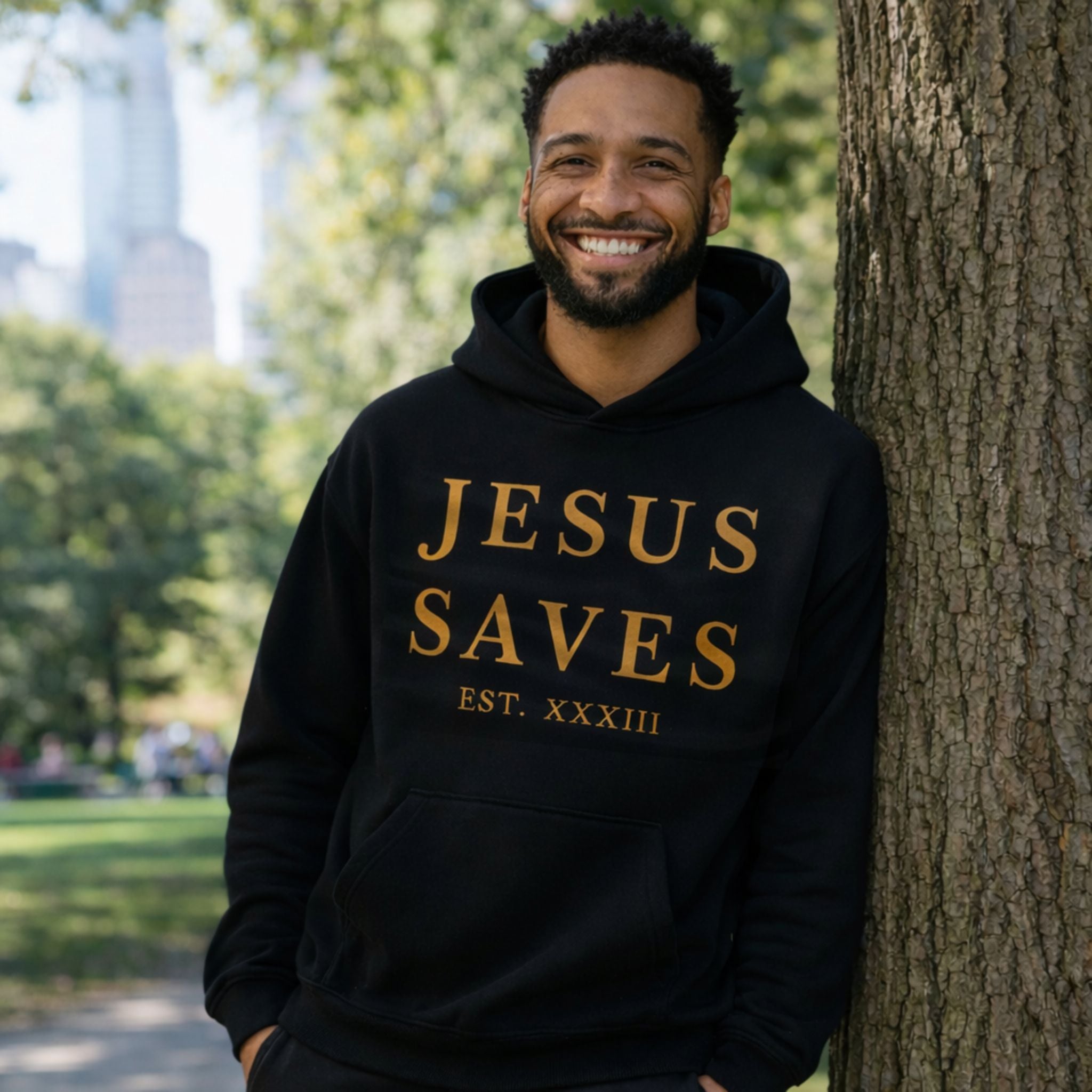 Man wearing a black hoodie with 'JESUS SAVES' text, standing next to a tree in a park.