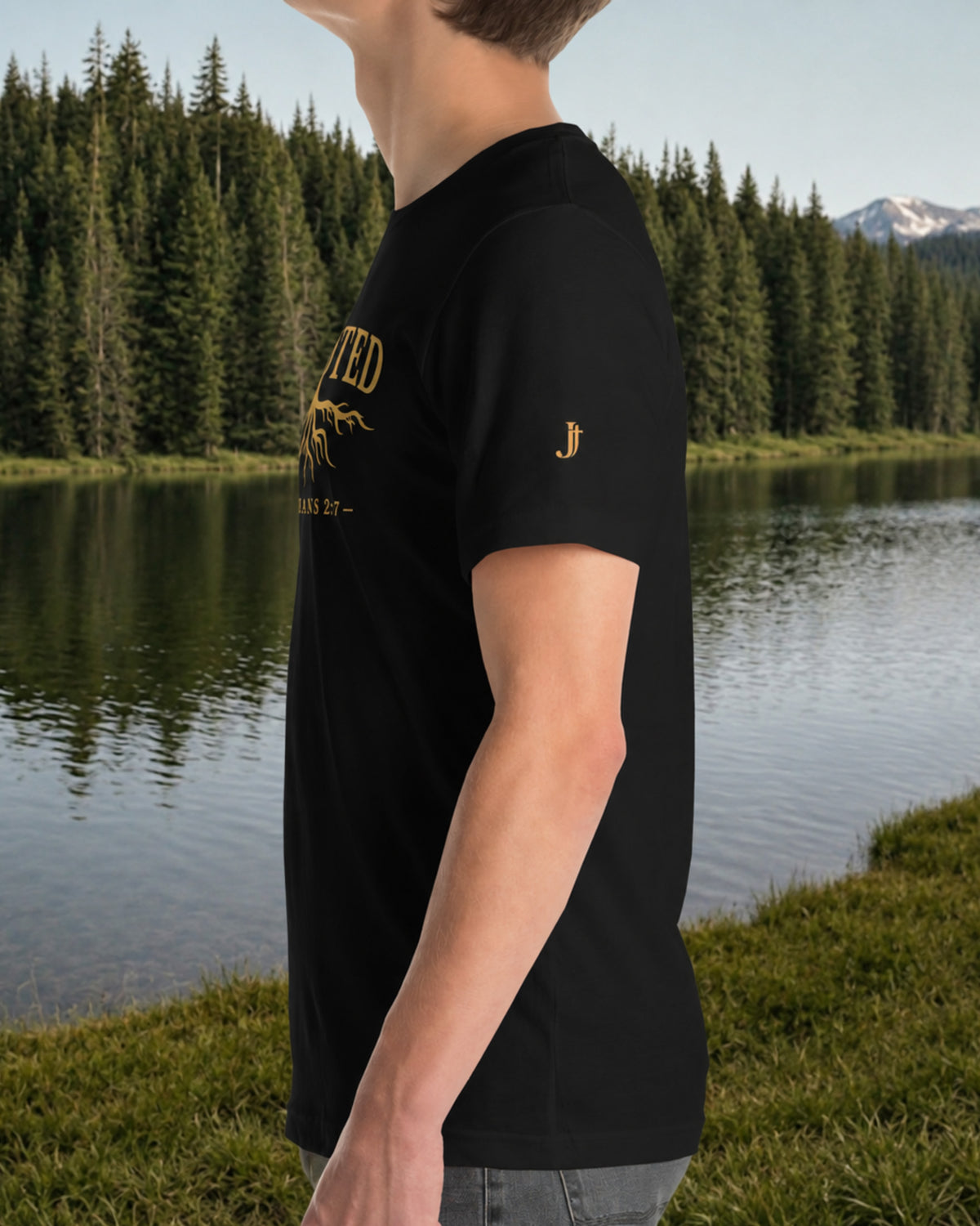A person wearing a black short-sleeve graphic T-shirt stands outdoors beside a lake with pine trees and mountains in the background. The shirt has gold printed text and a small gold logo on the sleeve.