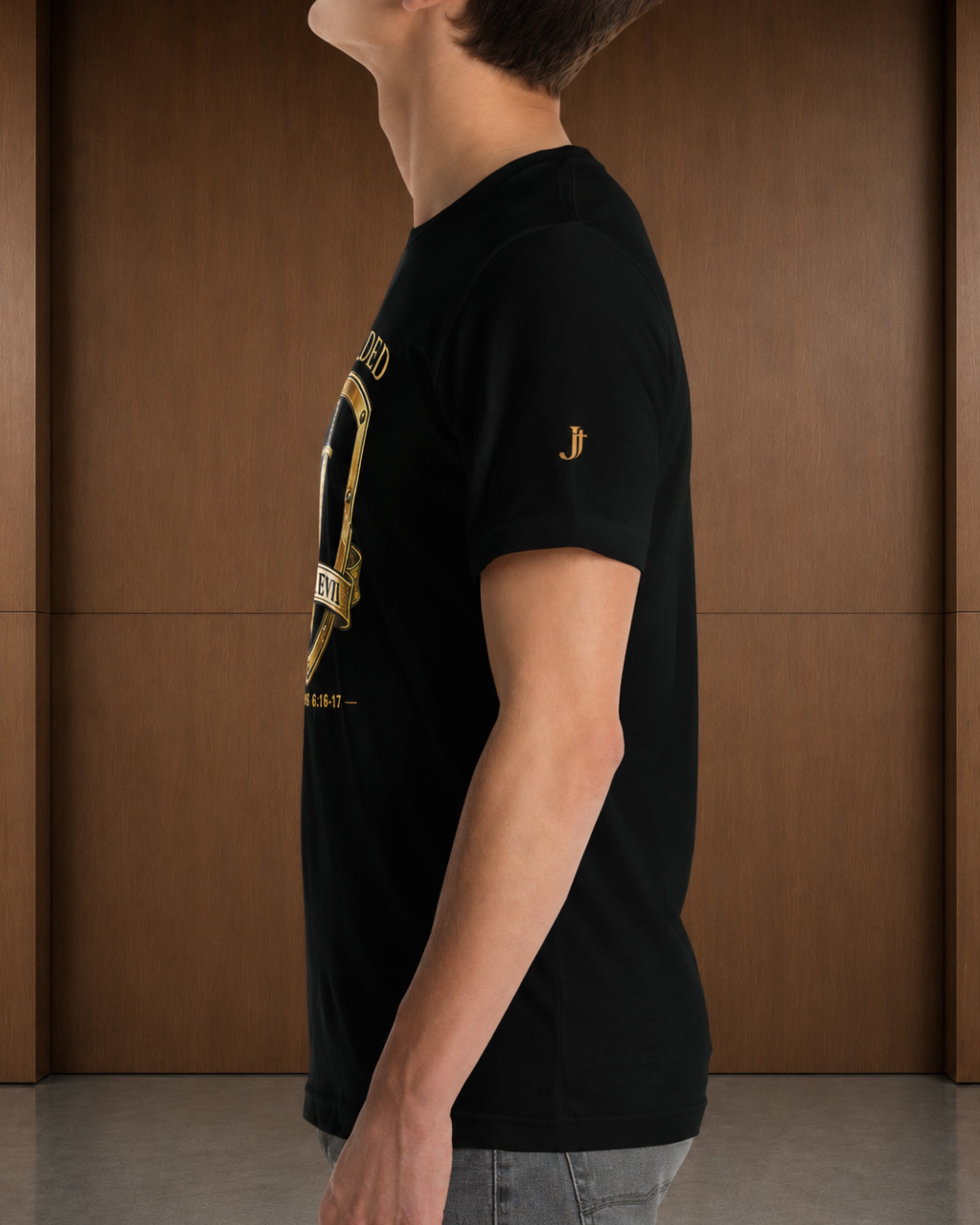 A man wearing a black short-sleeve t-shirt with a gold graphic print on the front and a small gold embroidered logo on the sleeve, standing in front of a brown wooden wall.