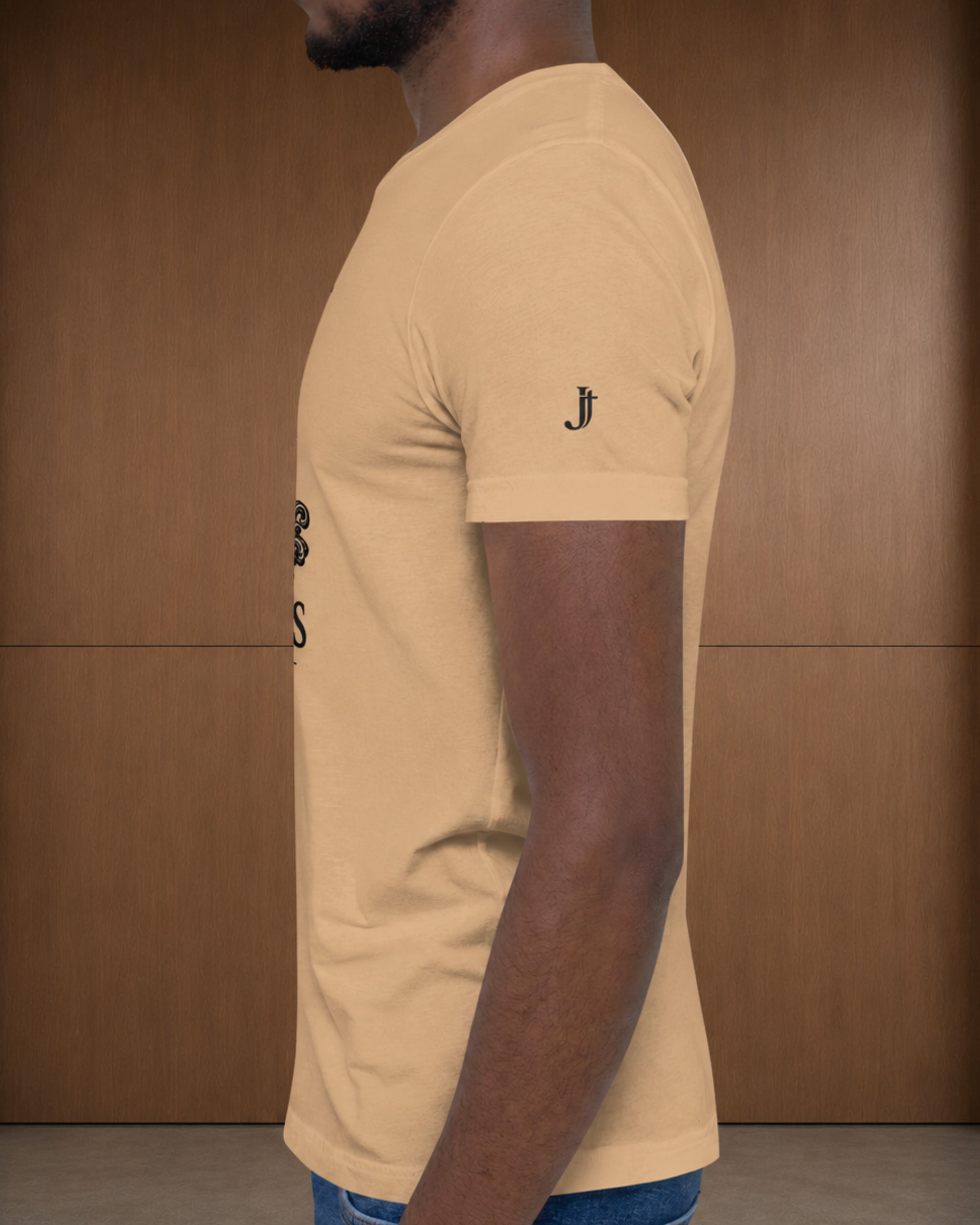 A man shown in profile wearing a light beige crew-neck t-shirt with a small black embroidered logo on the left sleeve, set against a brown wooden paneled background.