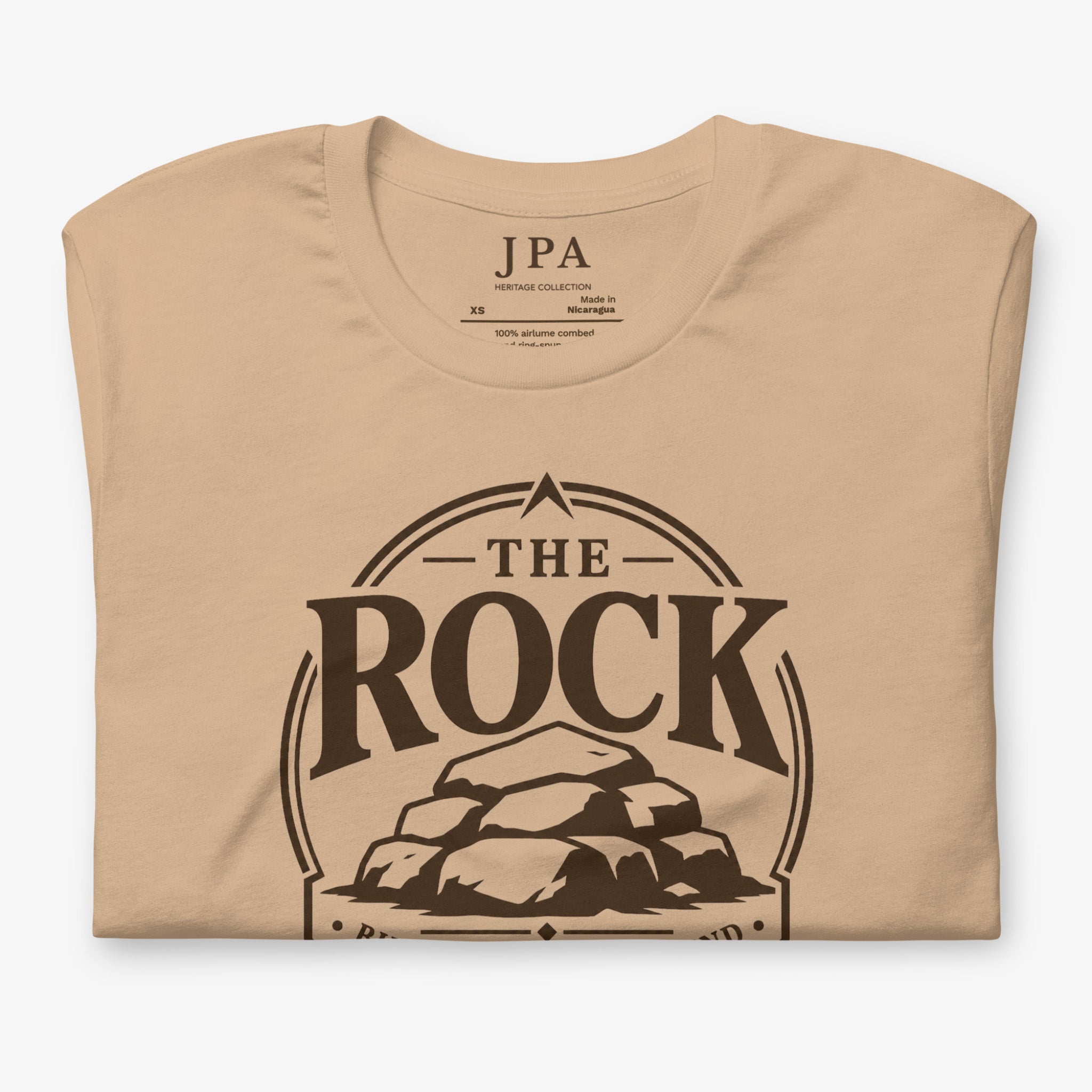A beige crewneck t-shirt with a dark brown graphic print reading 'THE ROCK' inside an oval border with a rock illustration, shown folded on a light gray background.