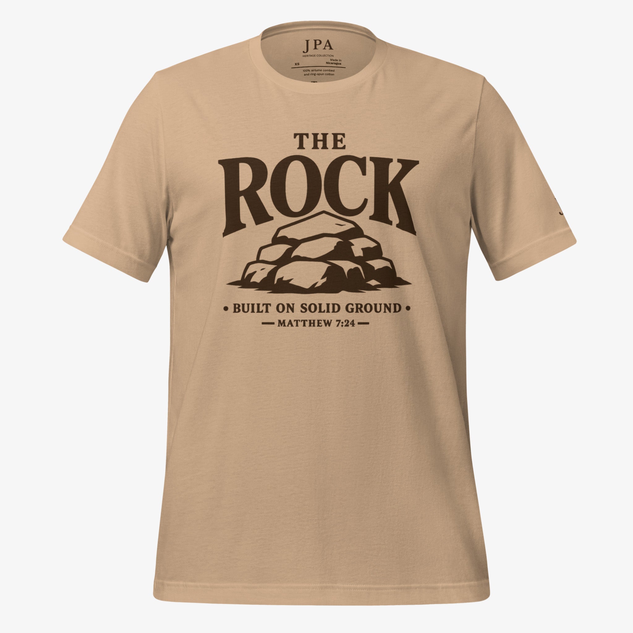 A beige crewneck t-shirt with a dark brown graphic print on the front reading 'THE ROCK BUILT ON SOLID GROUND MATTHEW 7:24' with an illustration of a rock formation. The brand 'JPA' is visible on the inner neck label.