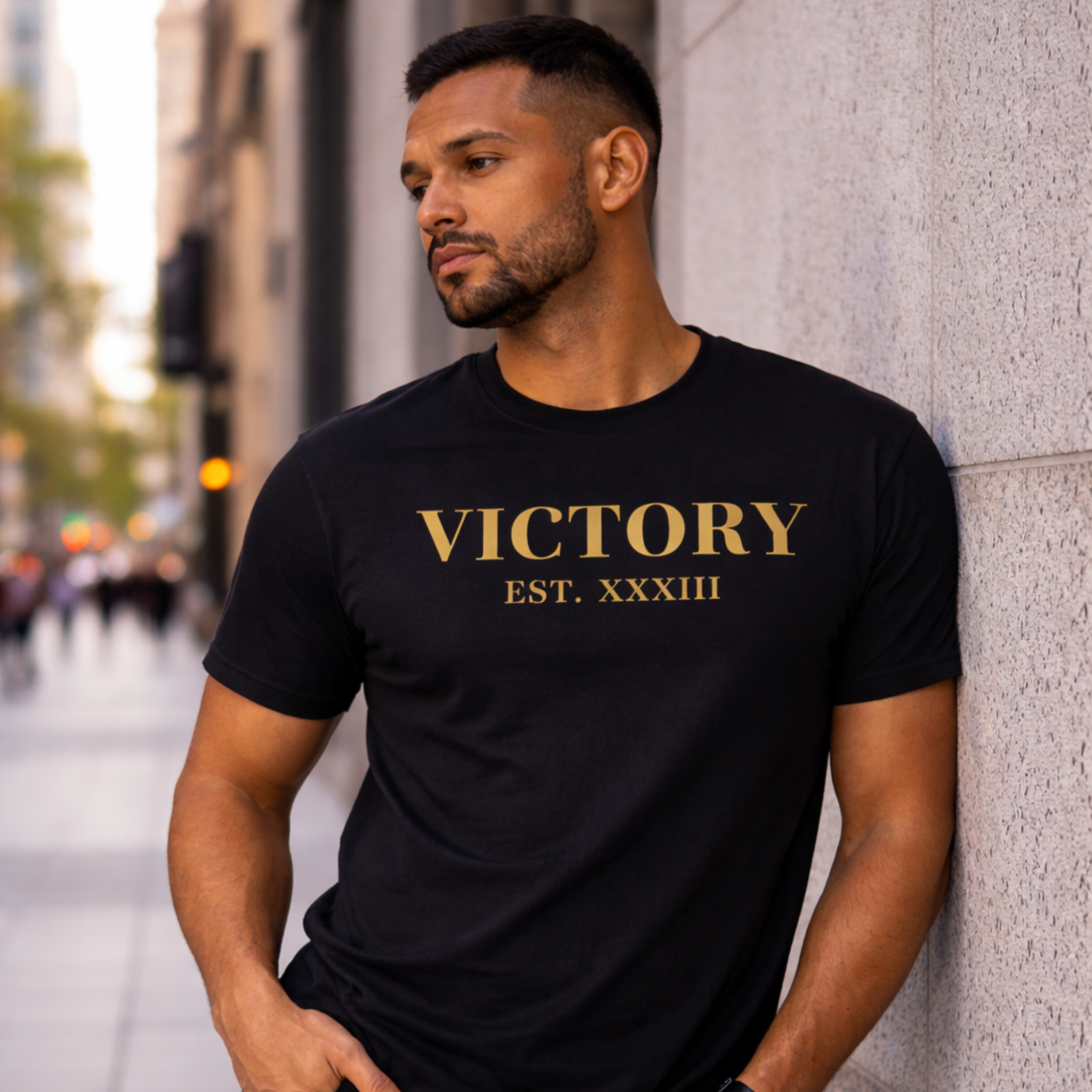 Man wearing a black t-shirt with 'VICTORY EST. XXXIII' in gold letters, standing against a city street background.
