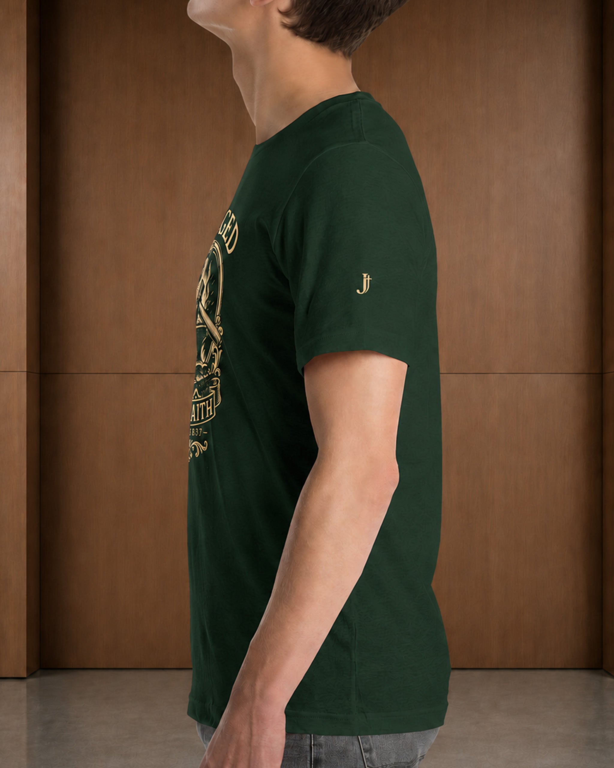A man wearing a dark forest green t-shirt with a gold graphic print on the front and a small gold logo on the sleeve, standing in front of a wooden paneled wall.