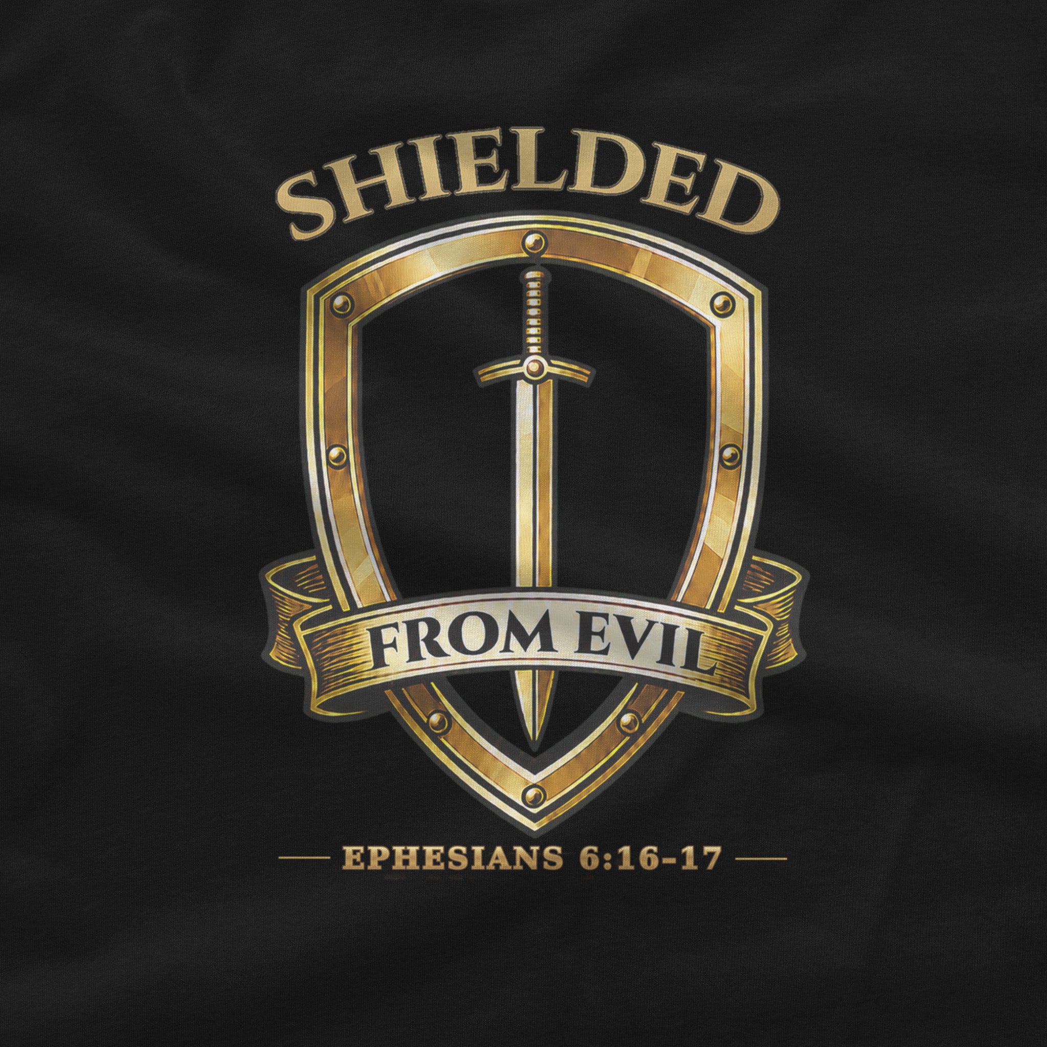 A close-up of a black fabric surface featuring a metallic gold graphic design. The design shows a shield with a sword inside, a ribbon banner reading 'FROM EVIL', and the word 'SHIELDED' above, with 'EPHESIANS 6:16-17' below.