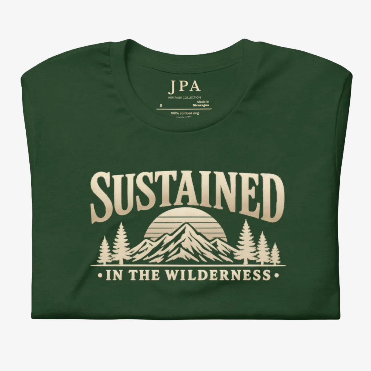 A dark forest green folded t-shirt with a beige graphic print that reads 'SUSTAINED IN THE WILDERNESS' above a mountain and pine tree illustration. The inside collar shows the brand name 'JPA'.