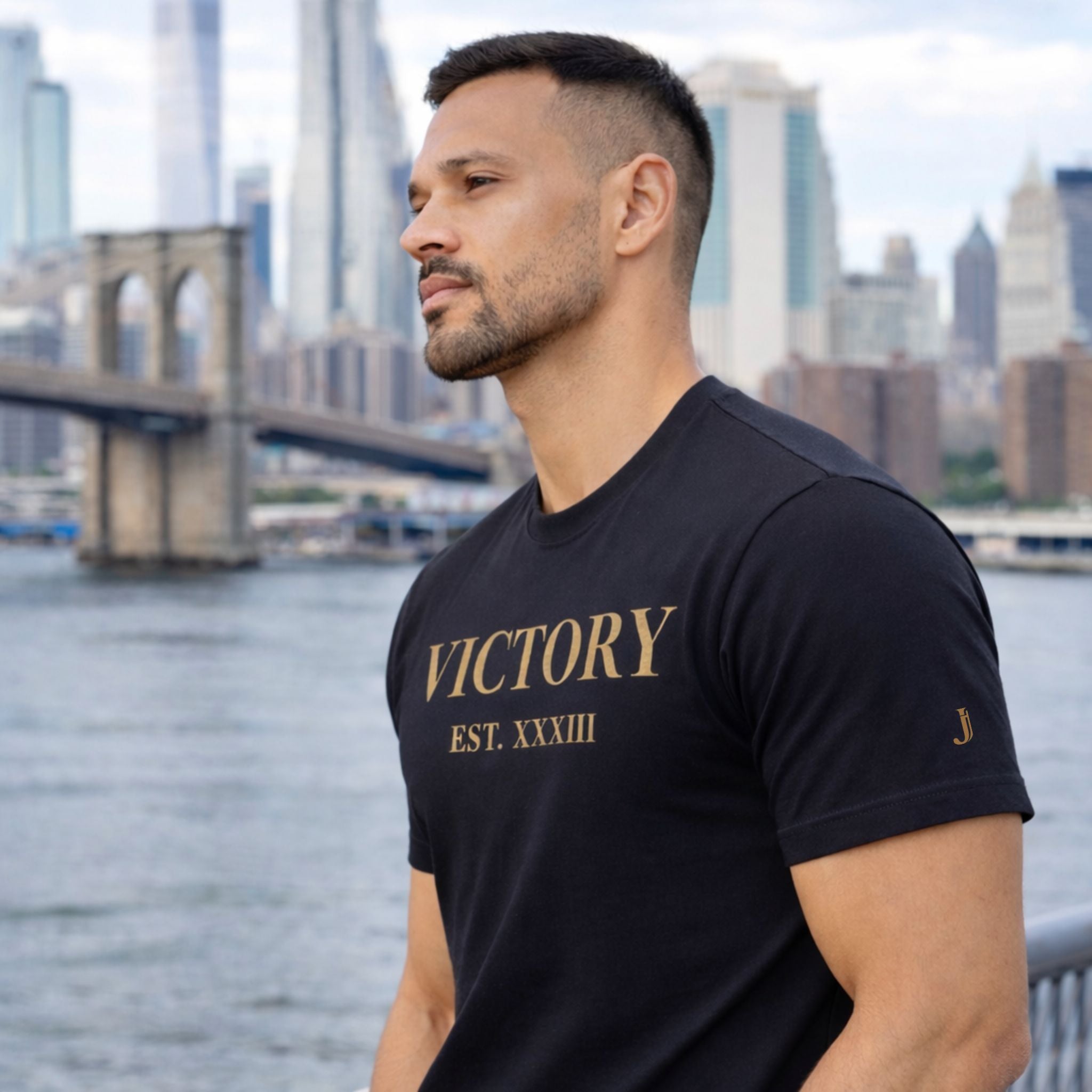 Man wearing a black t-shirt with 'VICTORY EST. XXXIII' text, standing by a waterfront with a city skyline in the background.