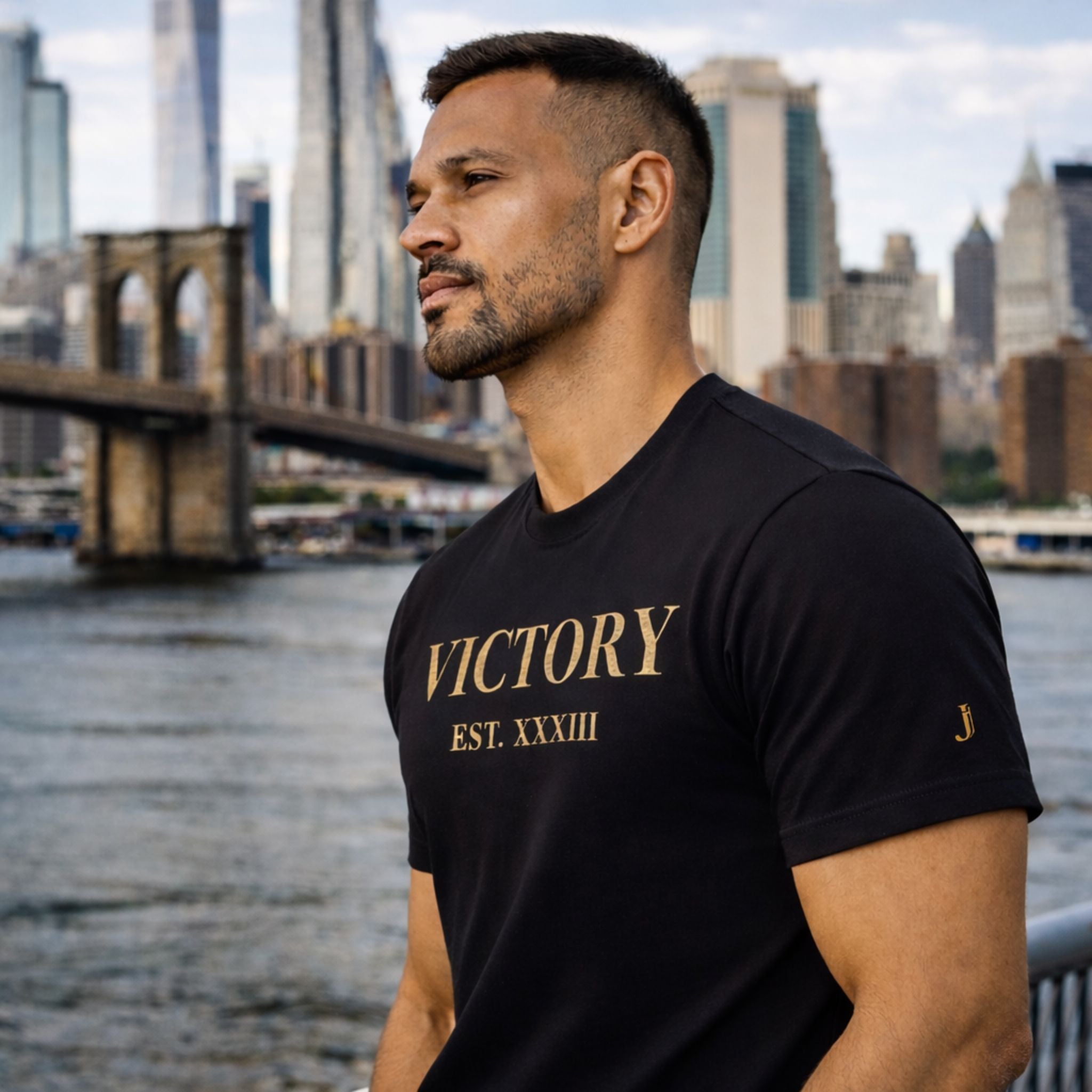 Man wearing a black t-shirt with 'VICTORY EST. XXXIII' text, standing by a waterfront with a city skyline in the background.