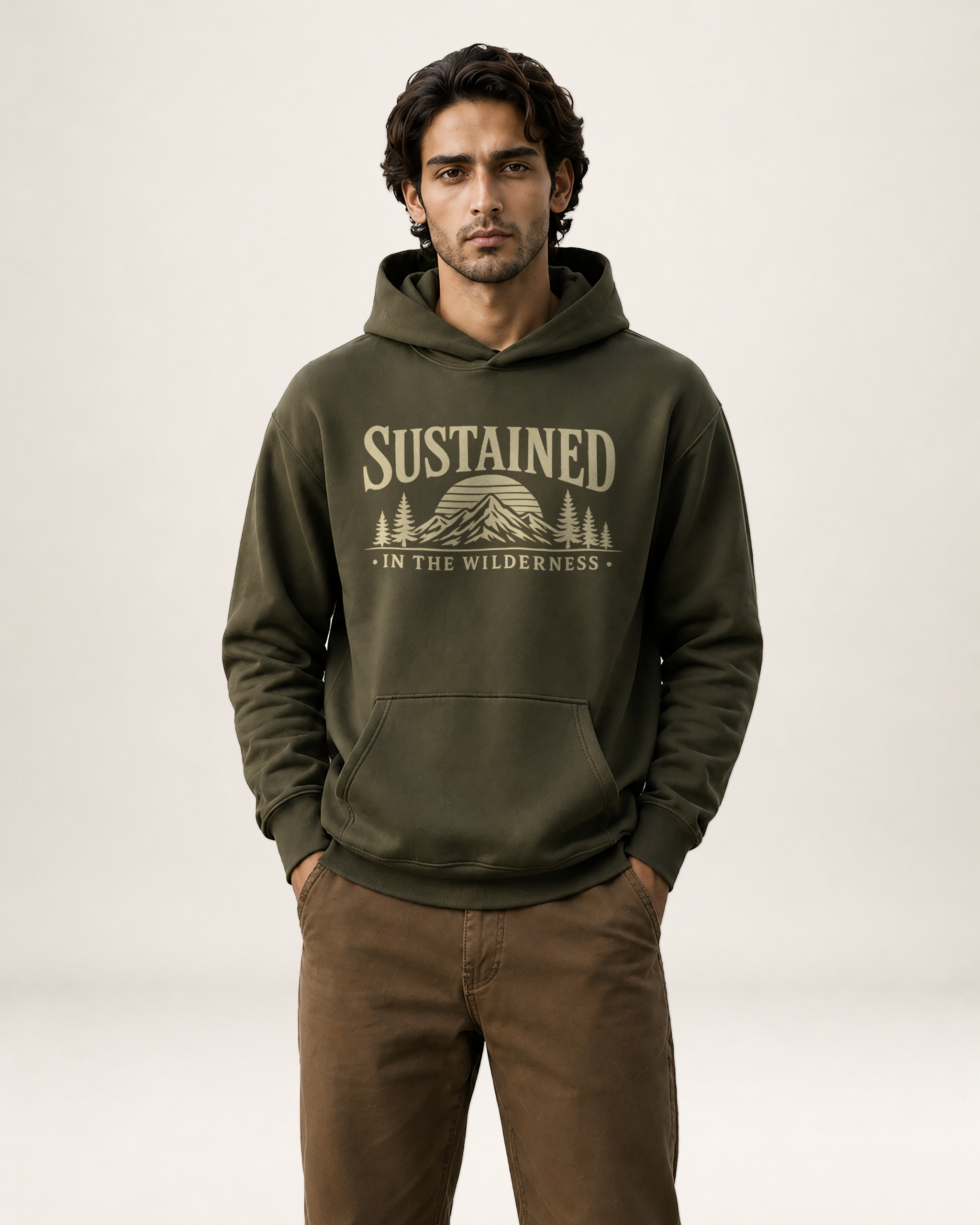 A man wearing an olive green pullover hoodie with a beige graphic print that reads 'SUSTAINED • IN THE WILDERNESS” above a mountain and tree illustration, paired with brown pants, against a plain light gray studio background.
