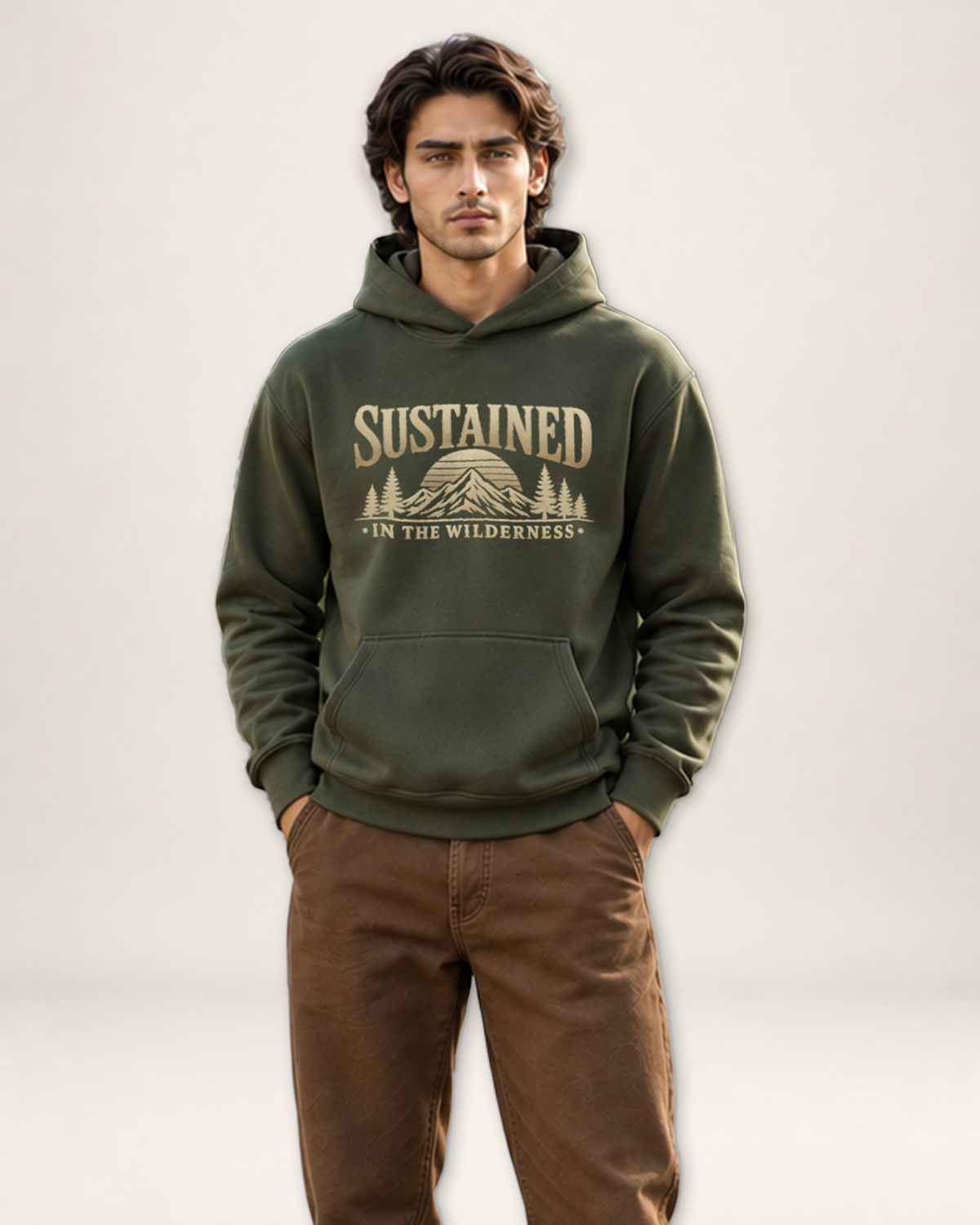 A man wearing an olive green pullover hoodie with a tan graphic print that reads 'SUSTAINED — IN THE WILDERNESS' above a mountain and trees, paired with brown pants, against a light gray studio background.