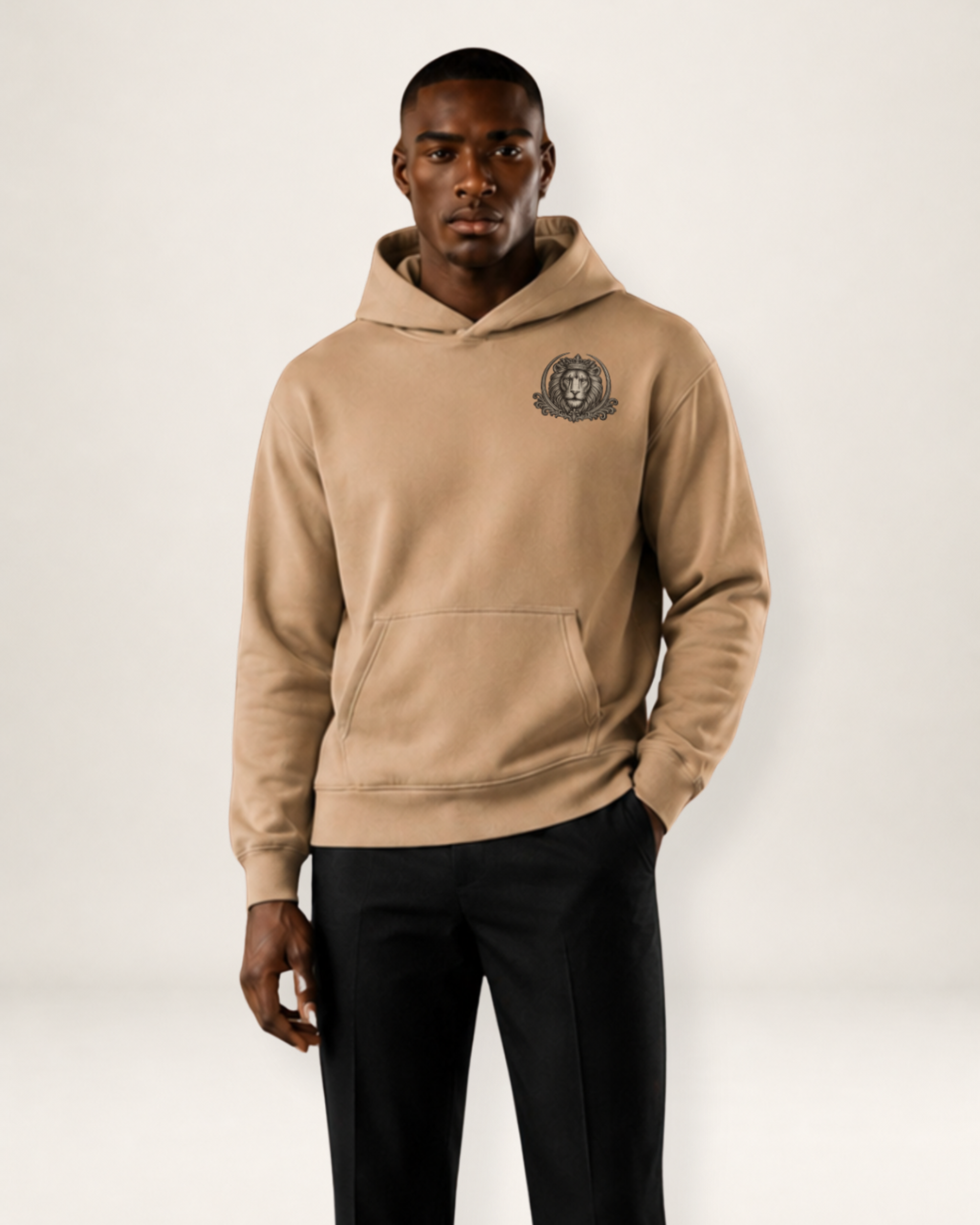 A male model wearing a beige pullover hoodie with a dark embroidered crest on the left chest, paired with black trousers, against a plain light gray studio background.