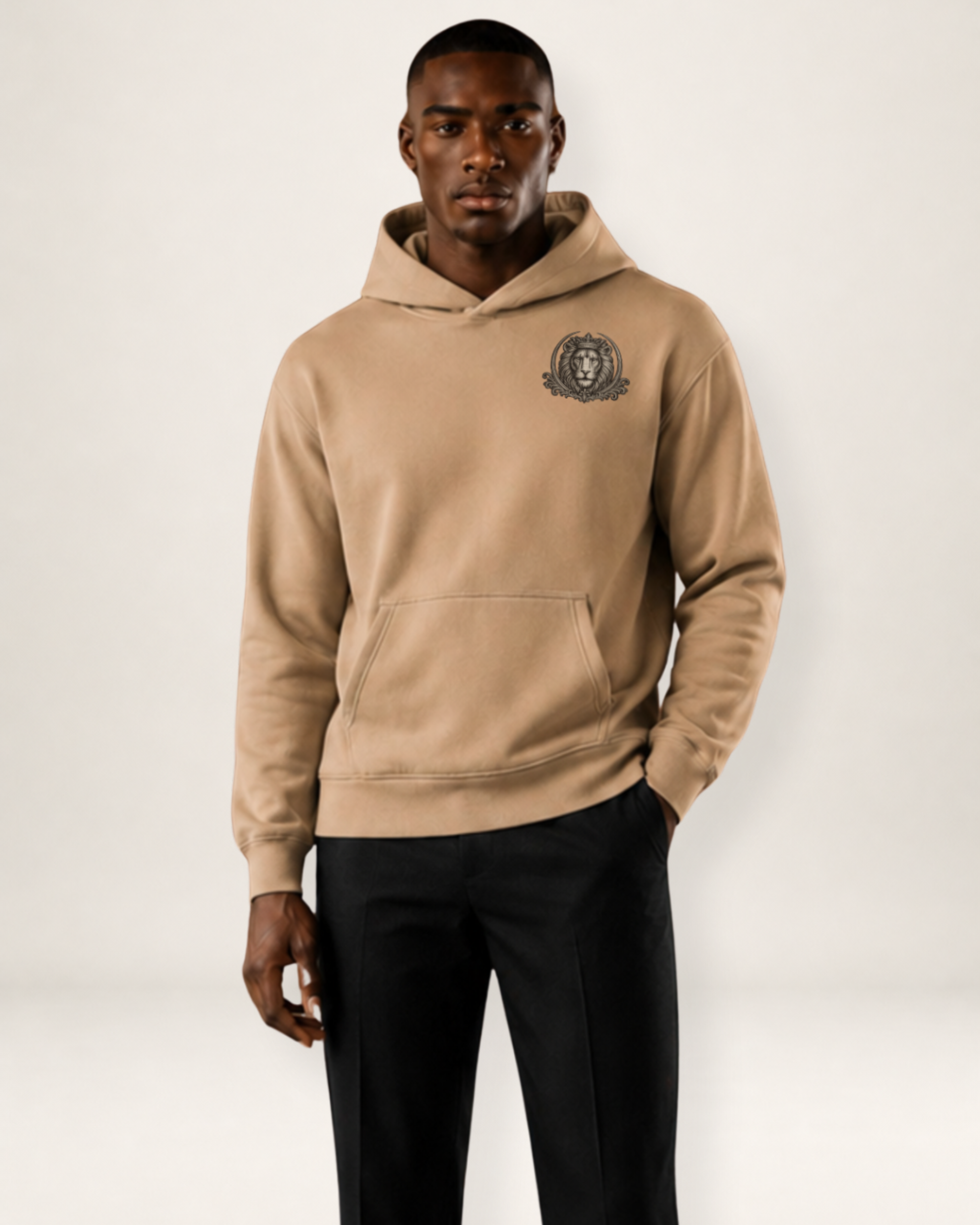 A man wearing a beige pullover hoodie with a dark gray graphic crest on the left chest, paired with black sweatpants, photographed against a plain light gray studio background.