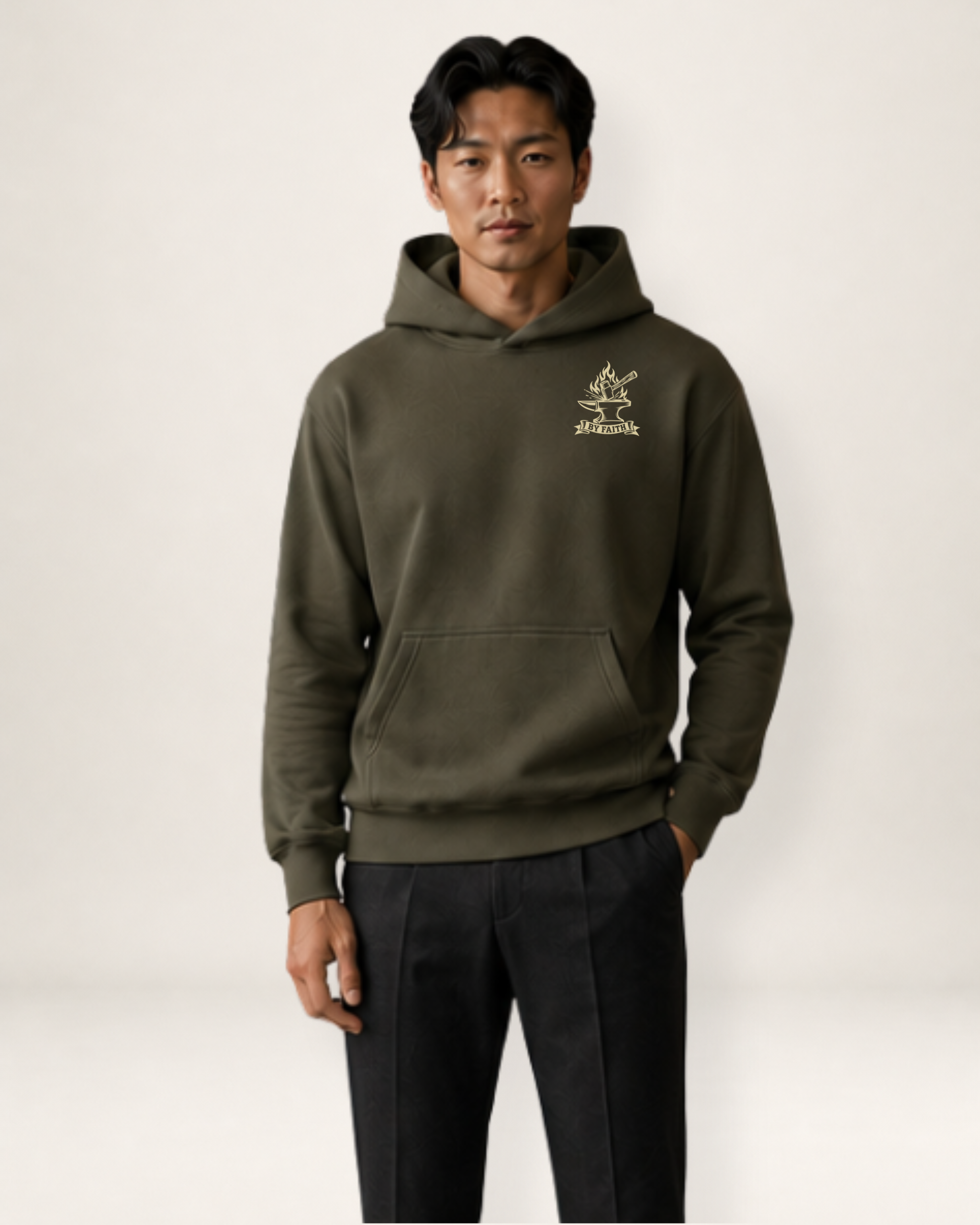 A man wearing an olive green pullover hoodie with a small gold embroidered logo on the left chest, paired with dark trousers, standing against a light gray studio background.