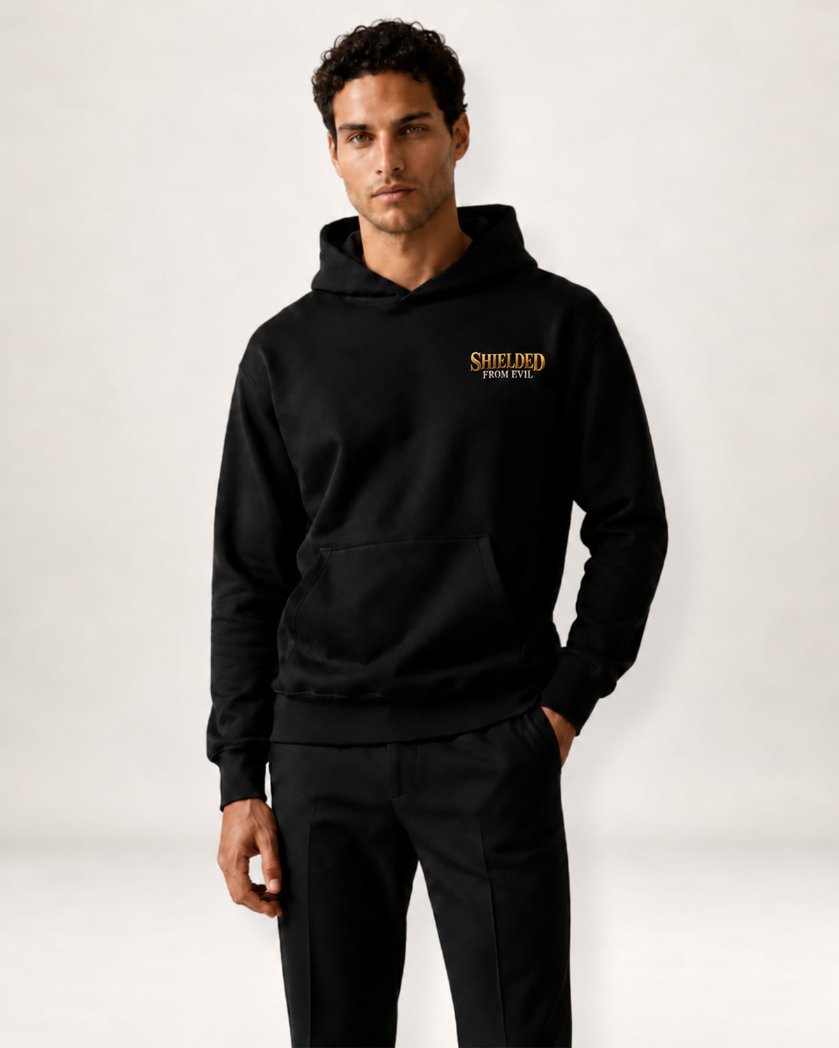 A man wearing a black pullover hoodie with a gold and black crest graphic on the left chest, paired with black sweatpants, photographed against a clean off-white studio background.
