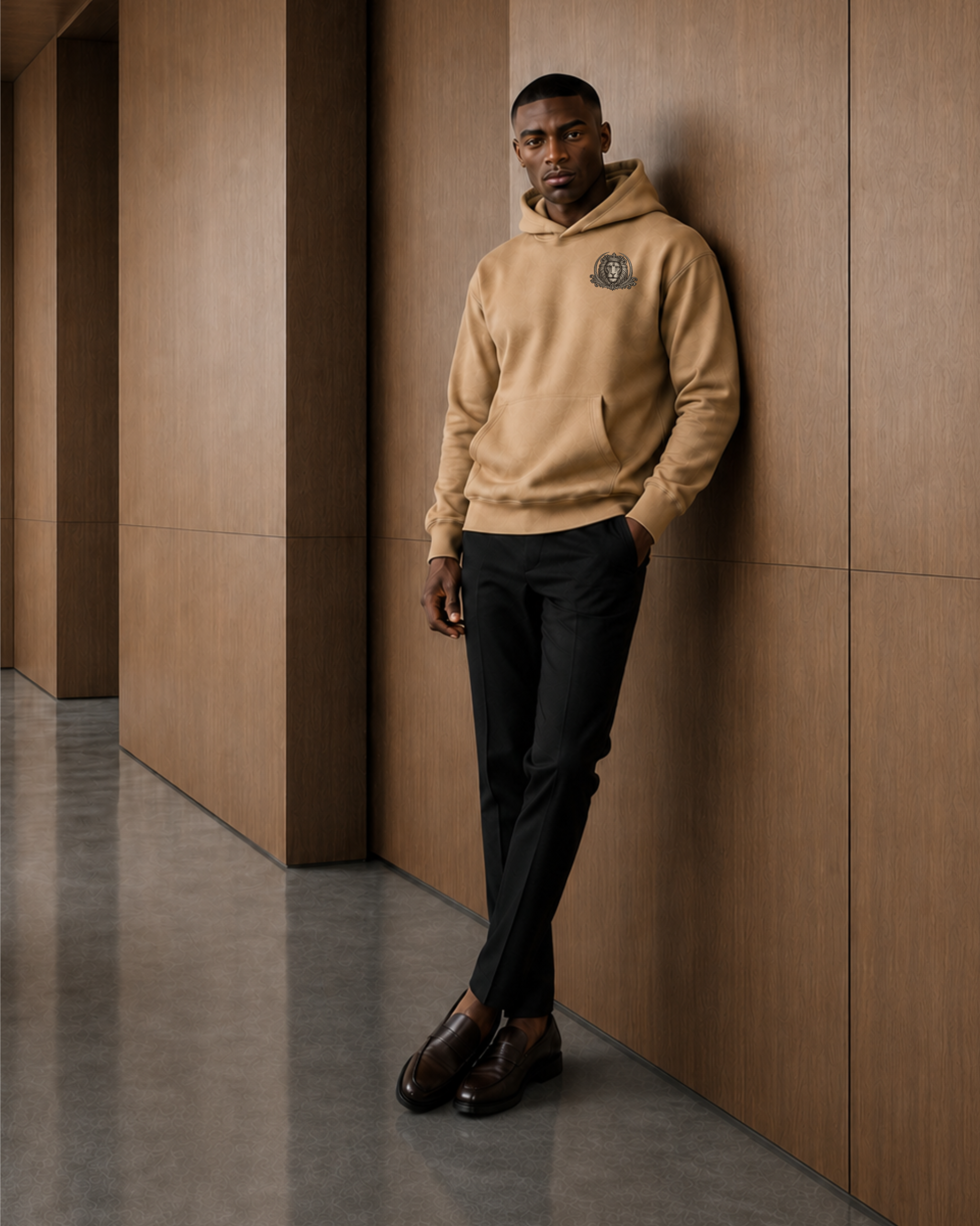A man stands against a wood-paneled wall wearing a tan hoodie with a small crest logo on the left chest, black trousers, and dark brown dress shoes in a modern interior hallway.