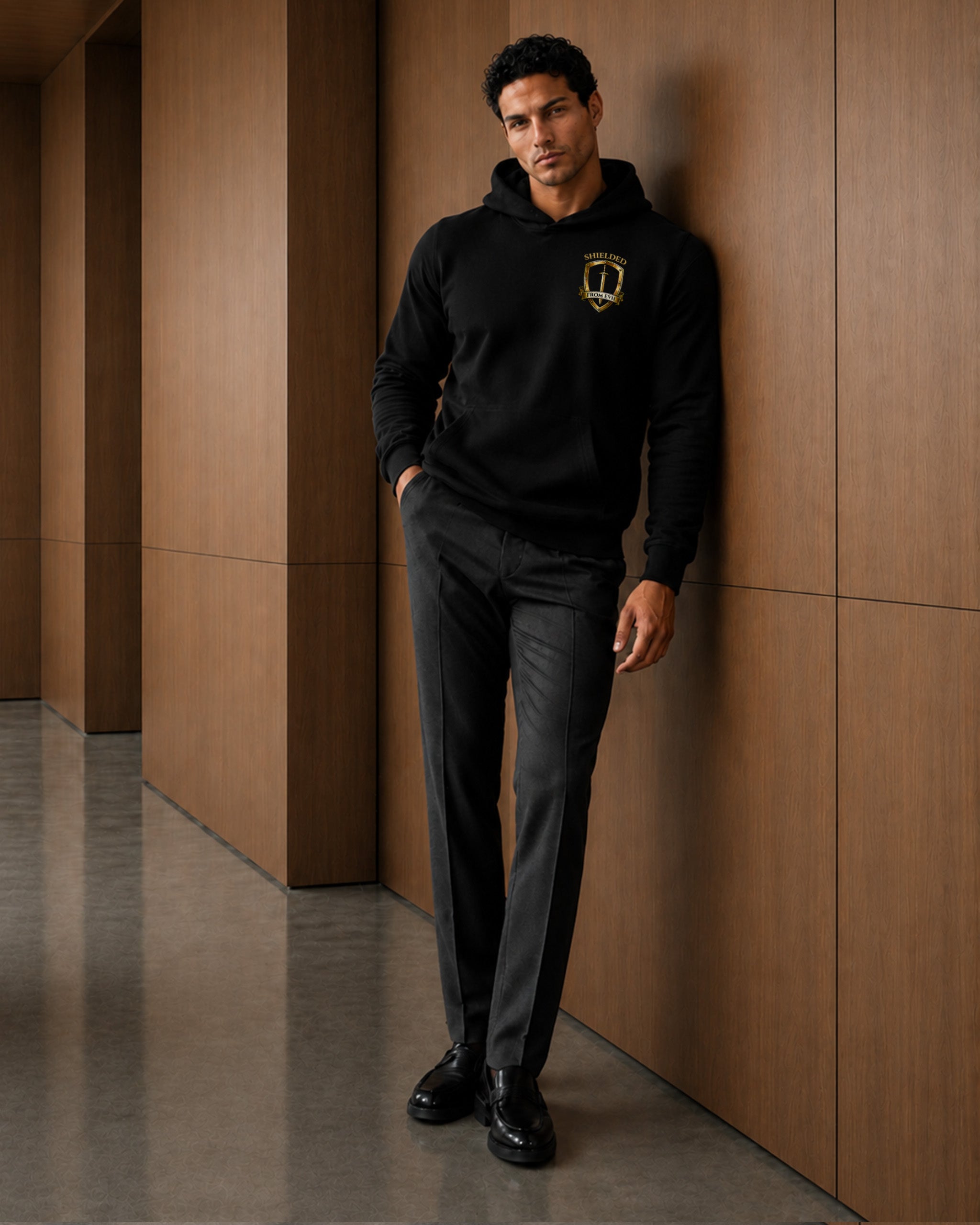A man wearing a black pullover hoodie with a gold crest logo on the left chest, paired with dark gray trousers and black dress shoes, leaning against a modern brown wood-paneled wall.