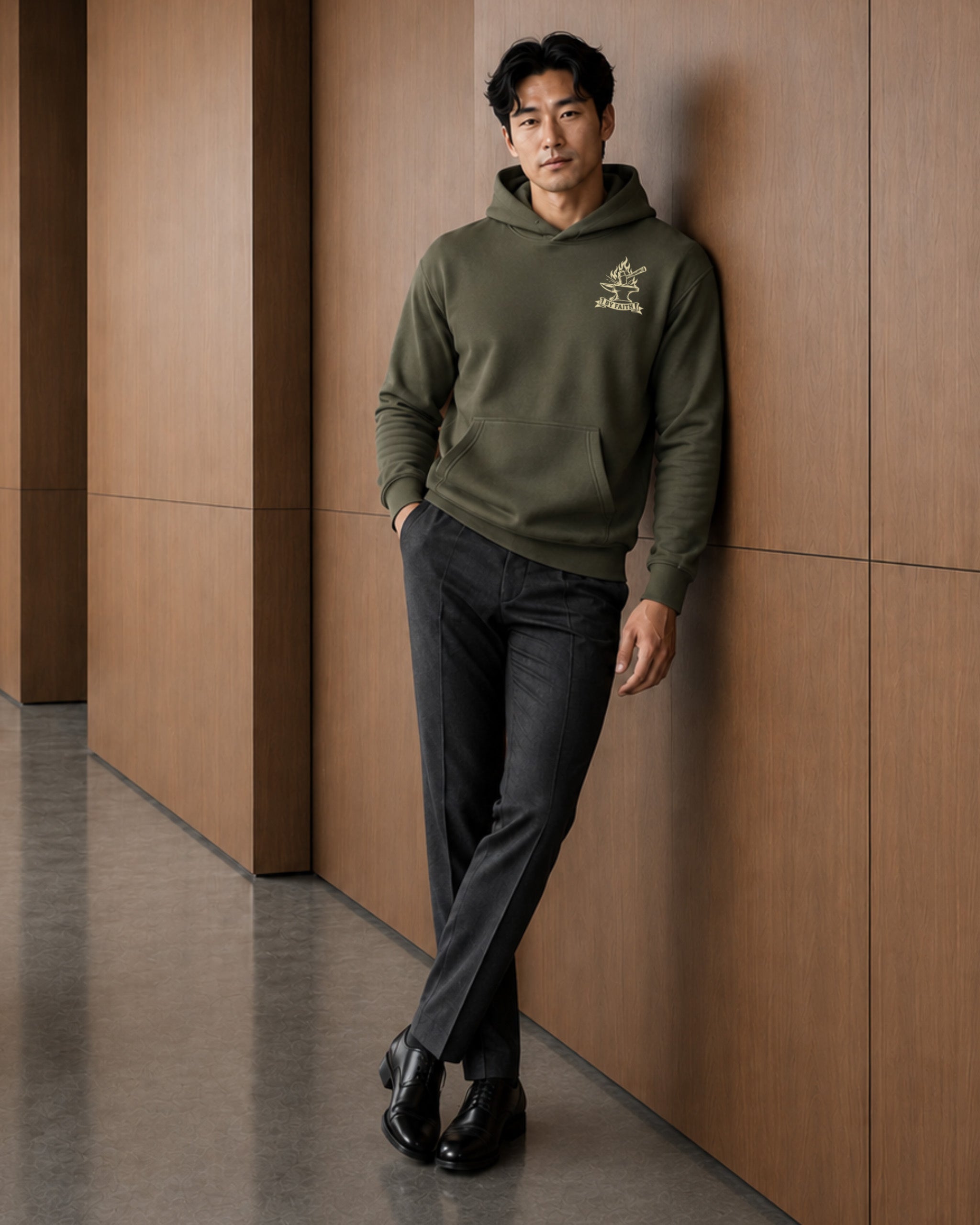 A man wearing an olive green hoodie with a gold embroidered logo on the left chest, paired with dark gray trousers and black shoes, leaning against a brown wood-paneled wall.