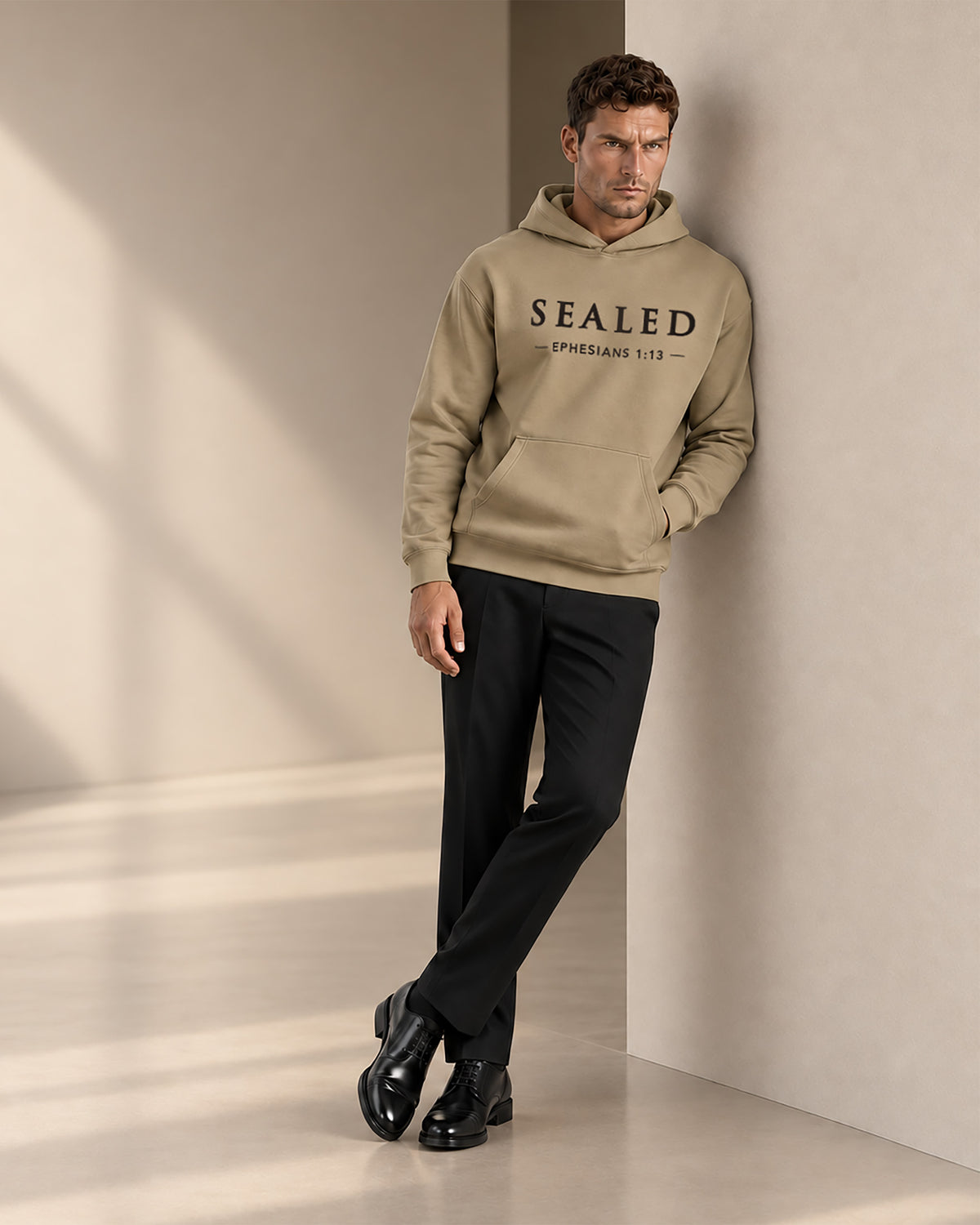 A man wearing a beige 'SEALED' branded hoodie and black trousers leans against a minimalist beige wall in a studio setting.