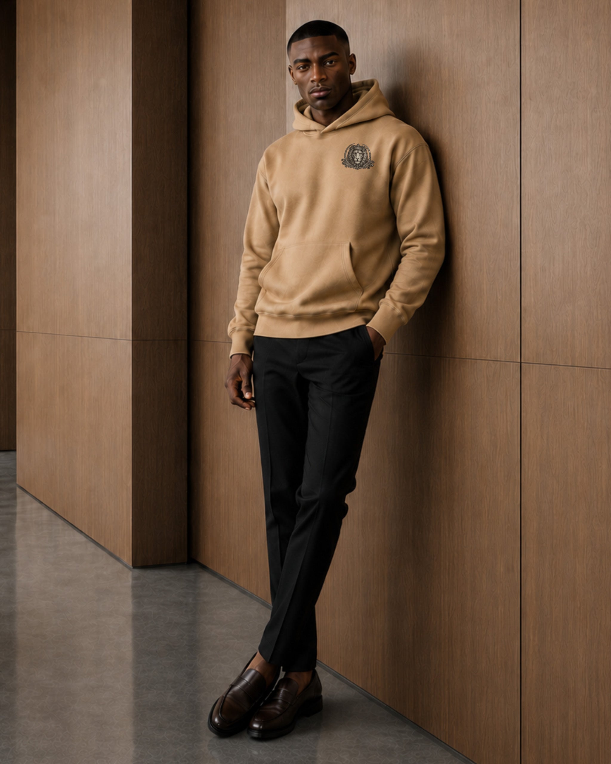 A man wearing a tan pullover hoodie with a small circular chest emblem, black trousers, and brown leather shoes, leaning against a brown wood-paneled wall.