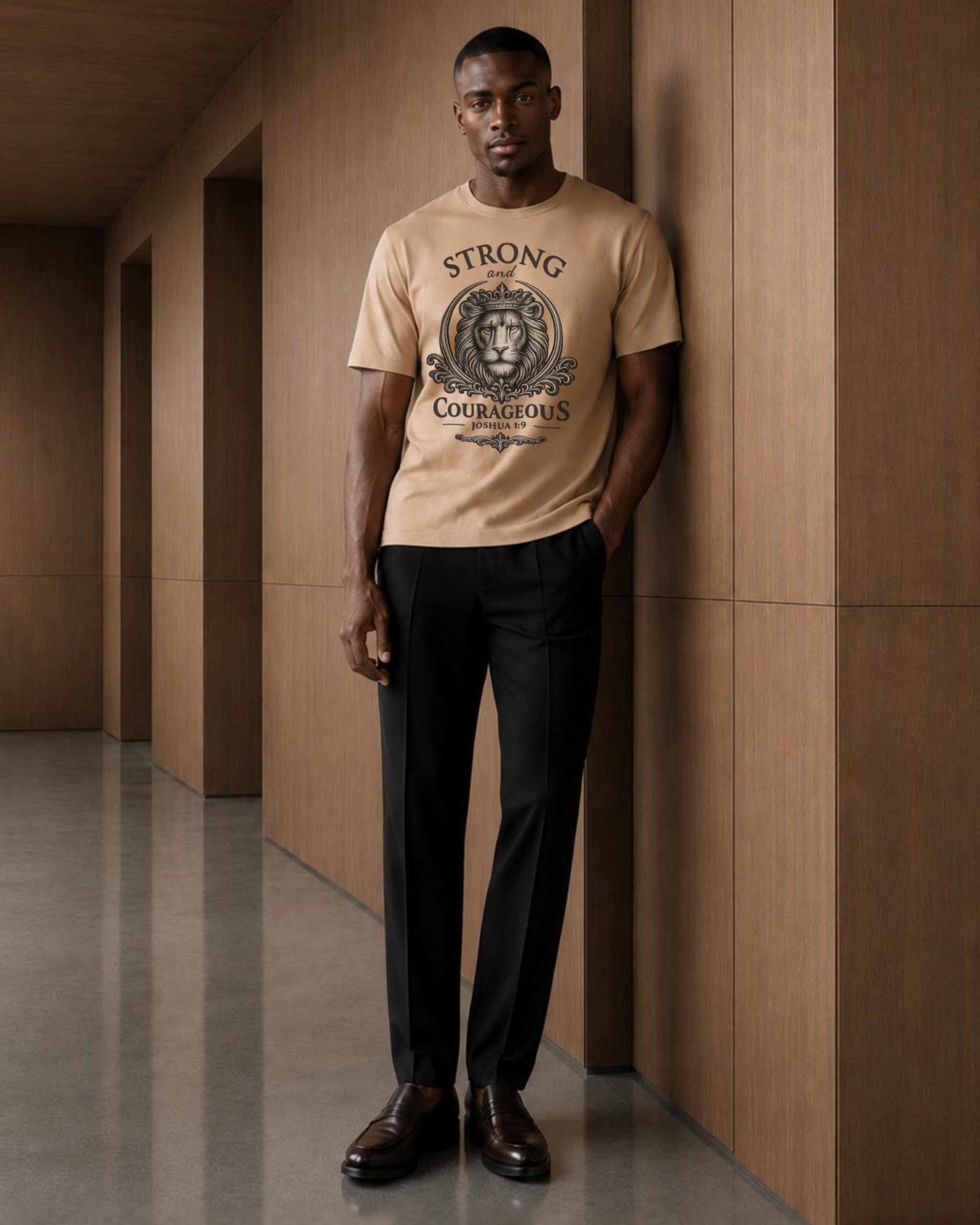 A man stands in a hallway wearing a beige graphic T-shirt with a lion crest and the words 'STRONG COURAGEOUS' printed on the front, paired with black trousers and brown leather shoes.