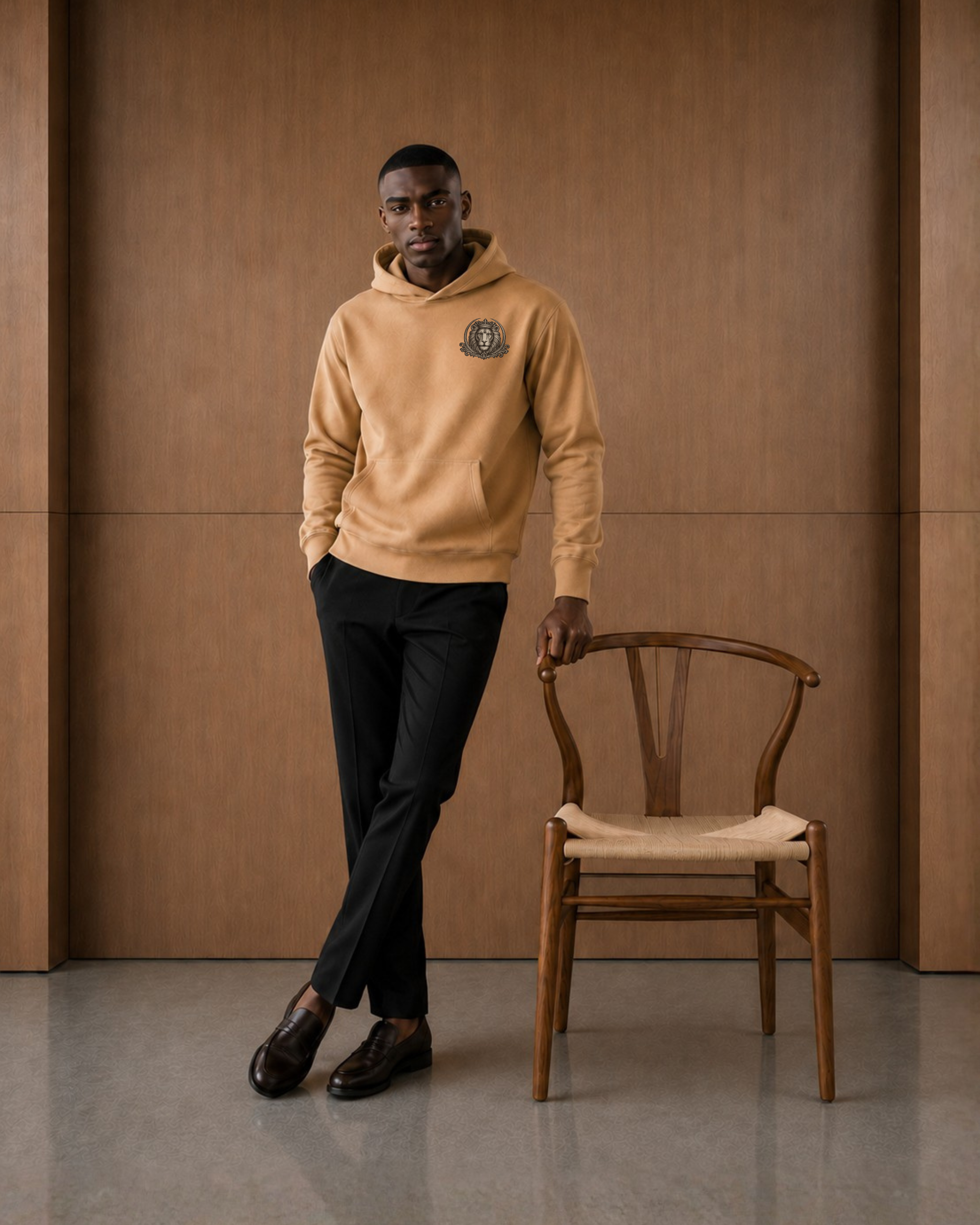 A man wearing a camel-colored hoodie with a crest logo on the left chest, black trousers, and brown leather shoes, standing next to a wooden chair in front of a wood-paneled wall.