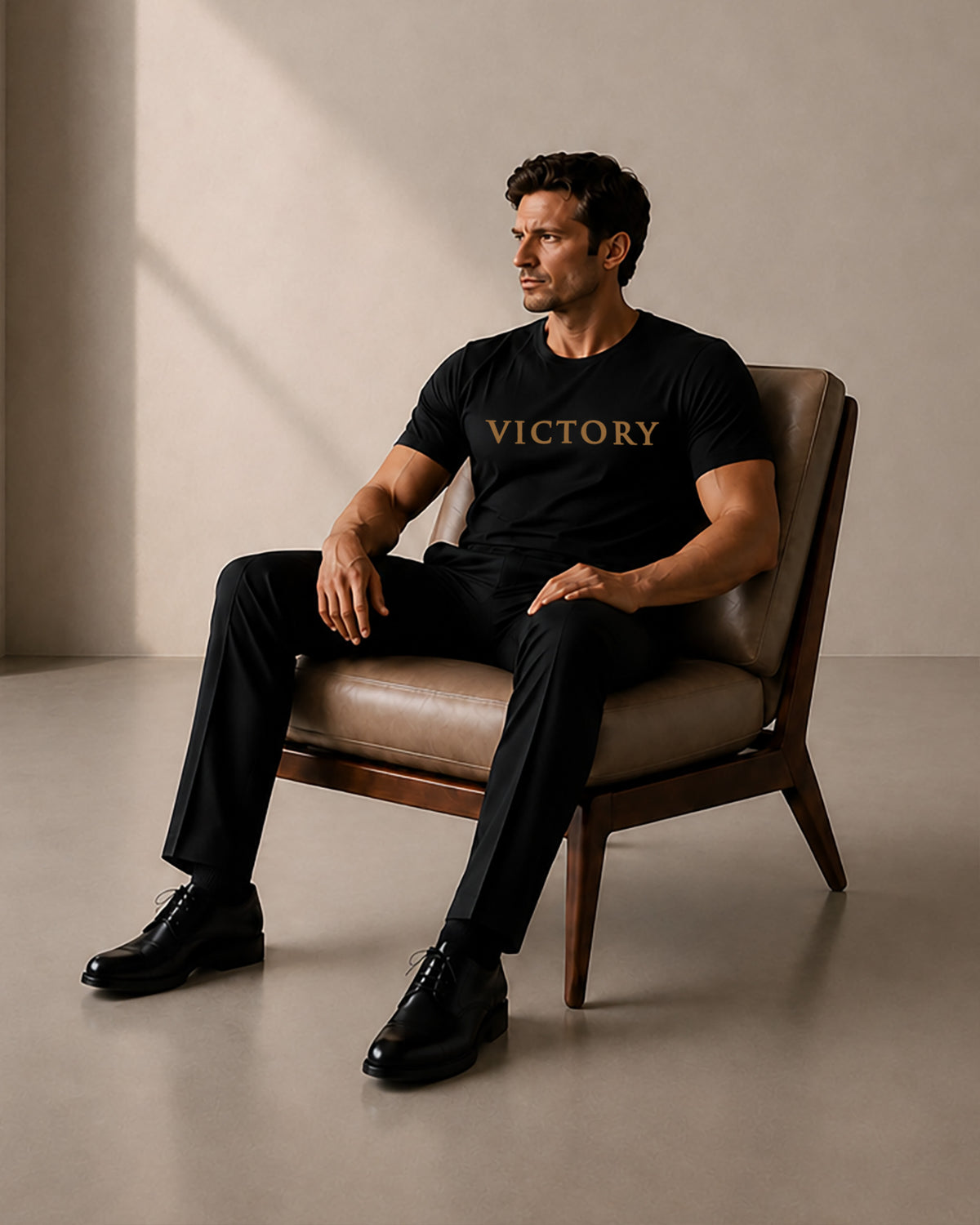 A man sits in a brown leather mid-century modern chair wearing a black t-shirt with 'VICTORY' printed in gold across the chest, paired with black trousers and black leather dress shoes, set against a minimalist beige studio background.