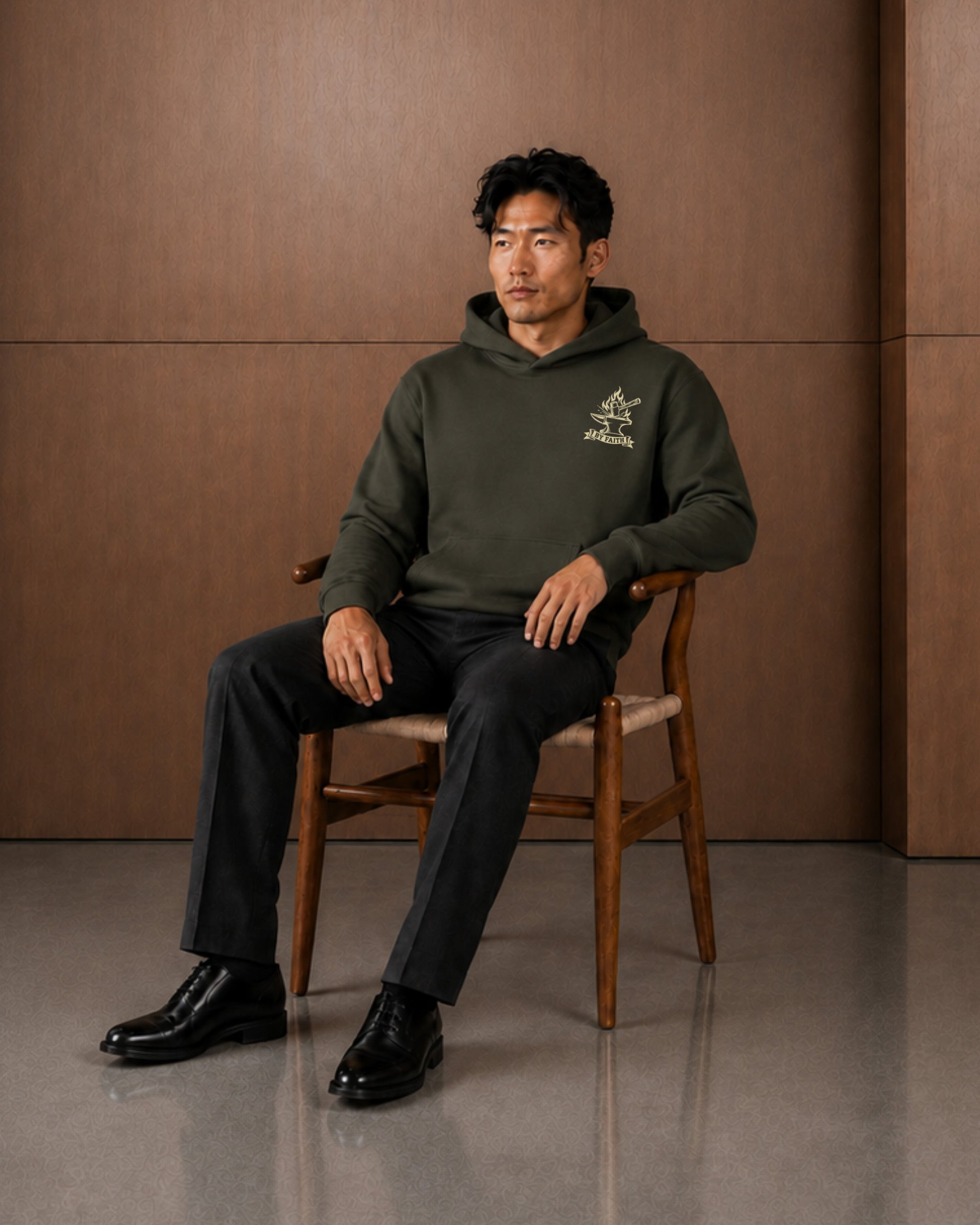 A man sits on a wooden chair wearing an olive green hooded sweatshirt with a small embroidered logo on the left chest, paired with black trousers and black dress shoes, against a warm brown paneled wall.