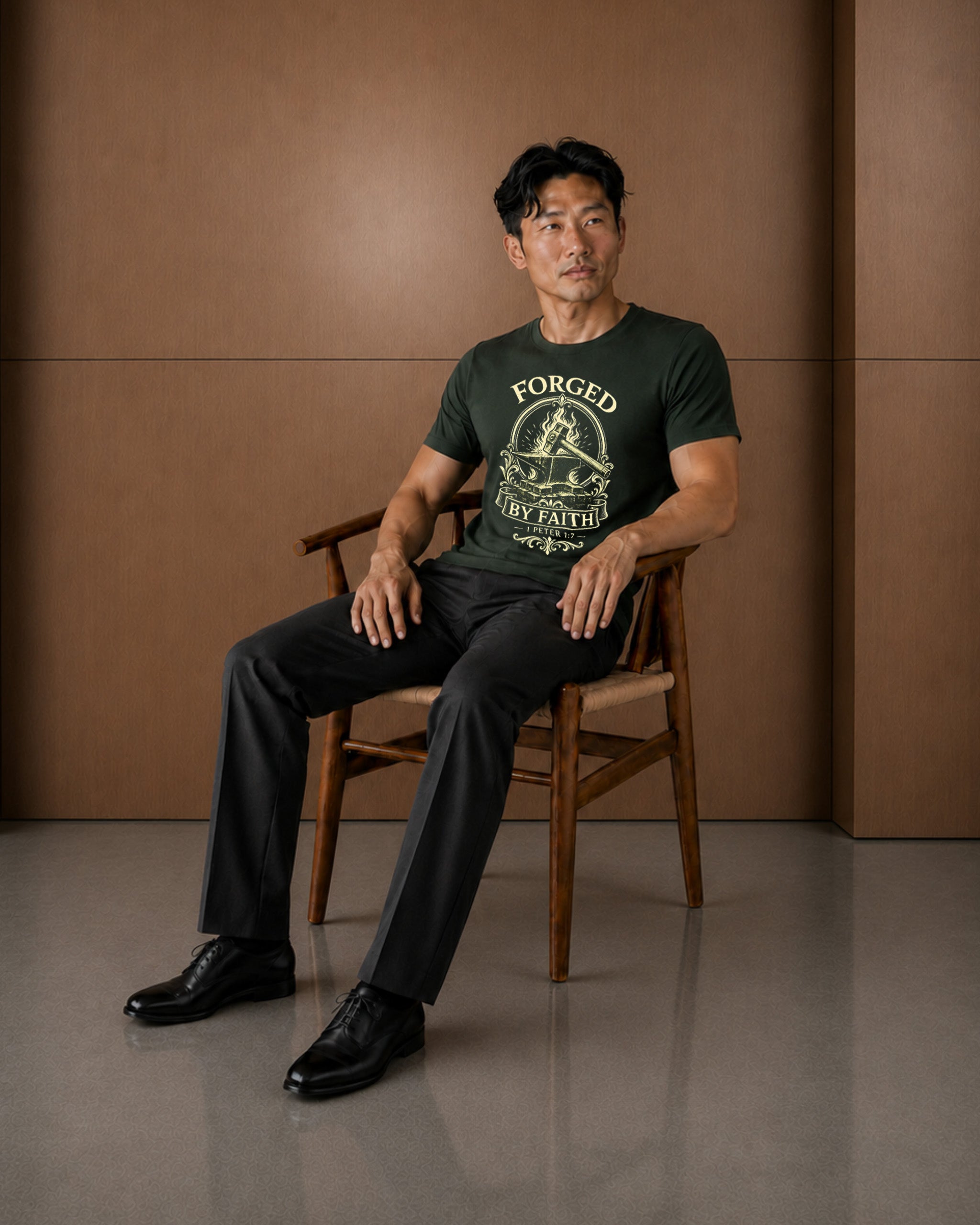 A man sits on a wooden chair wearing a dark green graphic t-shirt with 'FORGED IN FIRE' printed on the front, paired with black trousers and black leather shoes, set against a warm brown paneled wall.