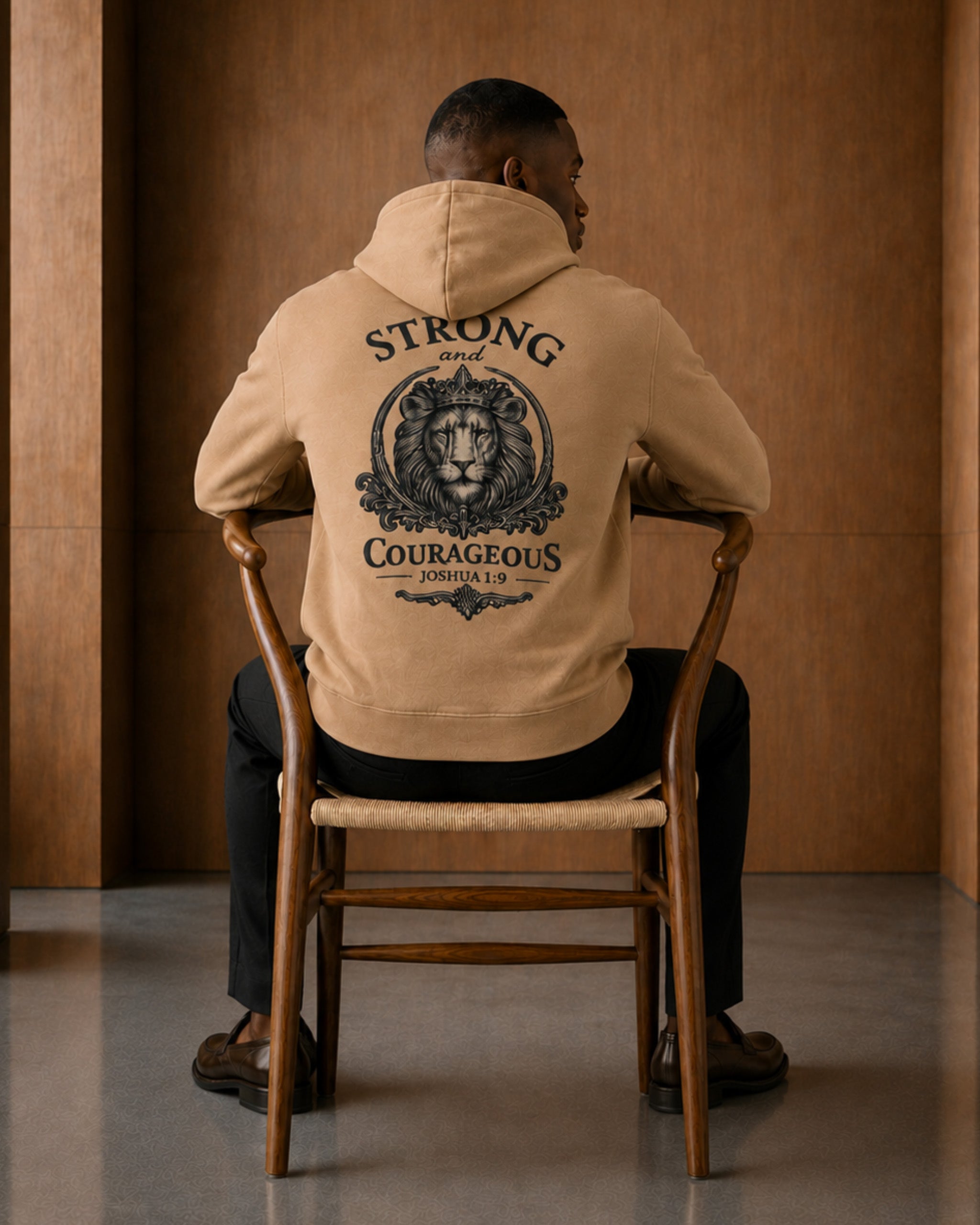 A man sits on a wooden chair facing away from the camera, wearing a tan hoodie with a large black graphic on the back featuring a lion and the words 'STRONG and COURAGEOUS'.