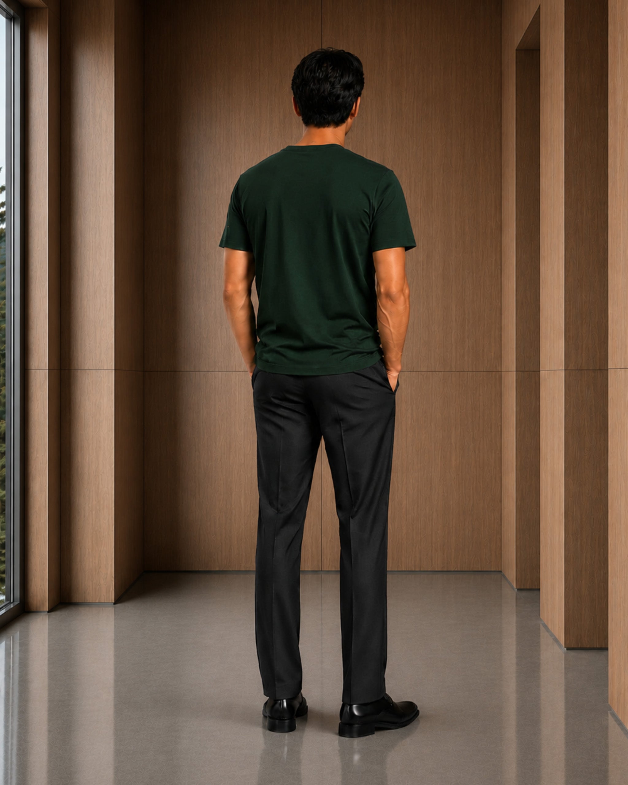 A man viewed from behind wearing a dark forest green short-sleeve t-shirt and black trousers, standing in a modern interior with wood-paneled walls and a polished floor.