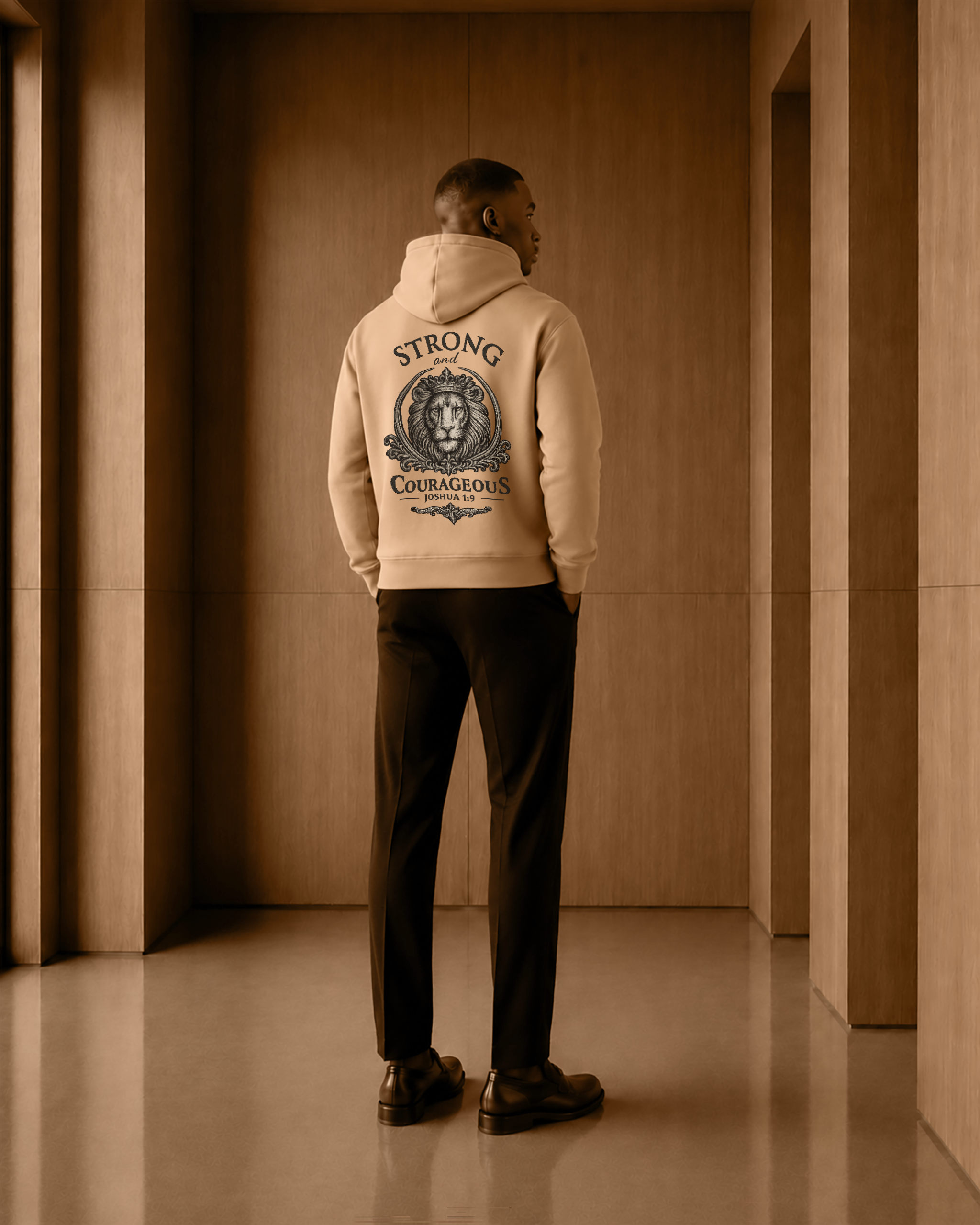 A man stands with his back to the camera wearing a beige hoodie with a large graphic print on the back, paired with dark trousers and brown shoes, in a warm-toned indoor hallway.