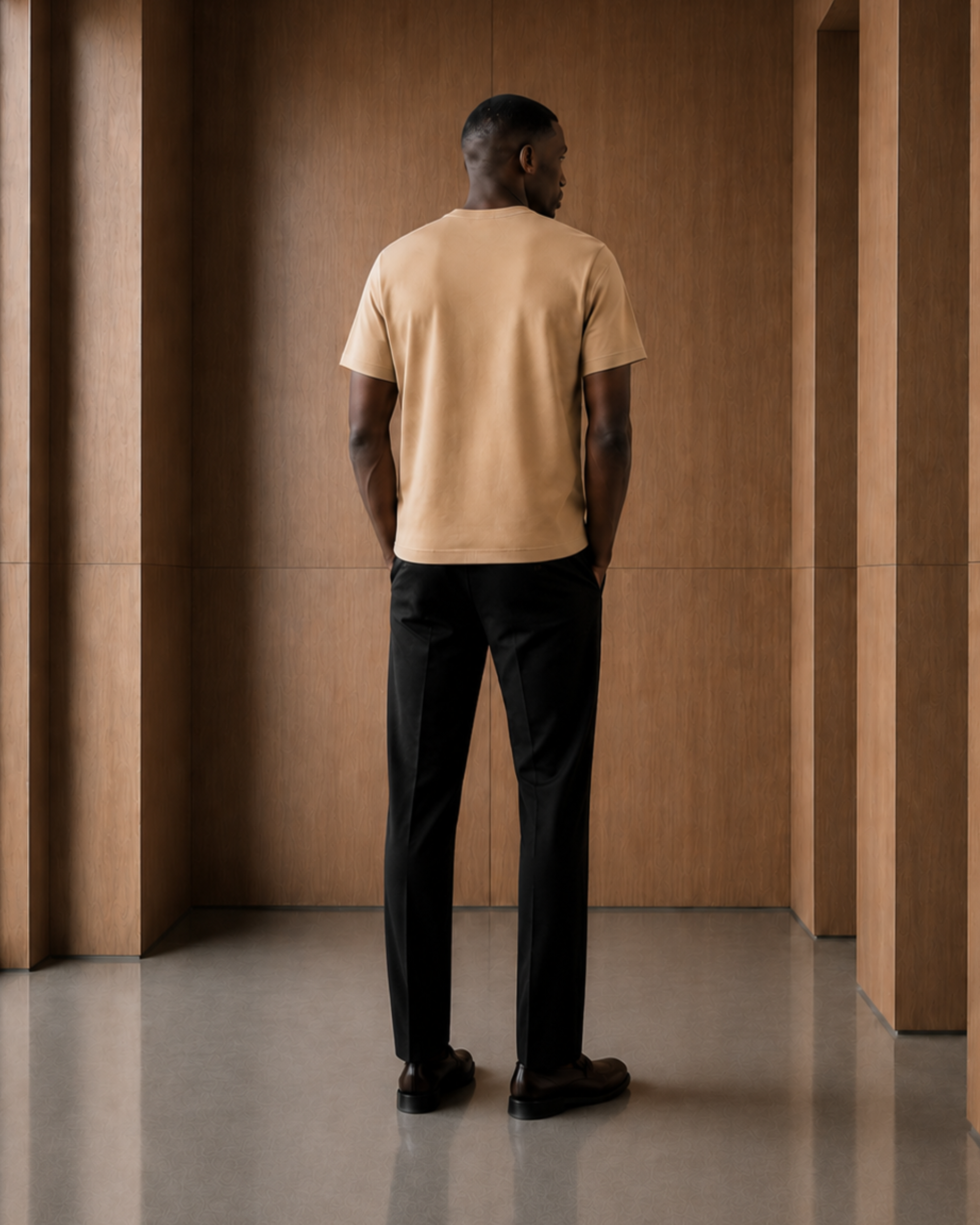 A man viewed from behind wearing a light tan crew-neck t-shirt and black trousers, standing in a modern interior with warm wood-paneled walls and a polished floor.