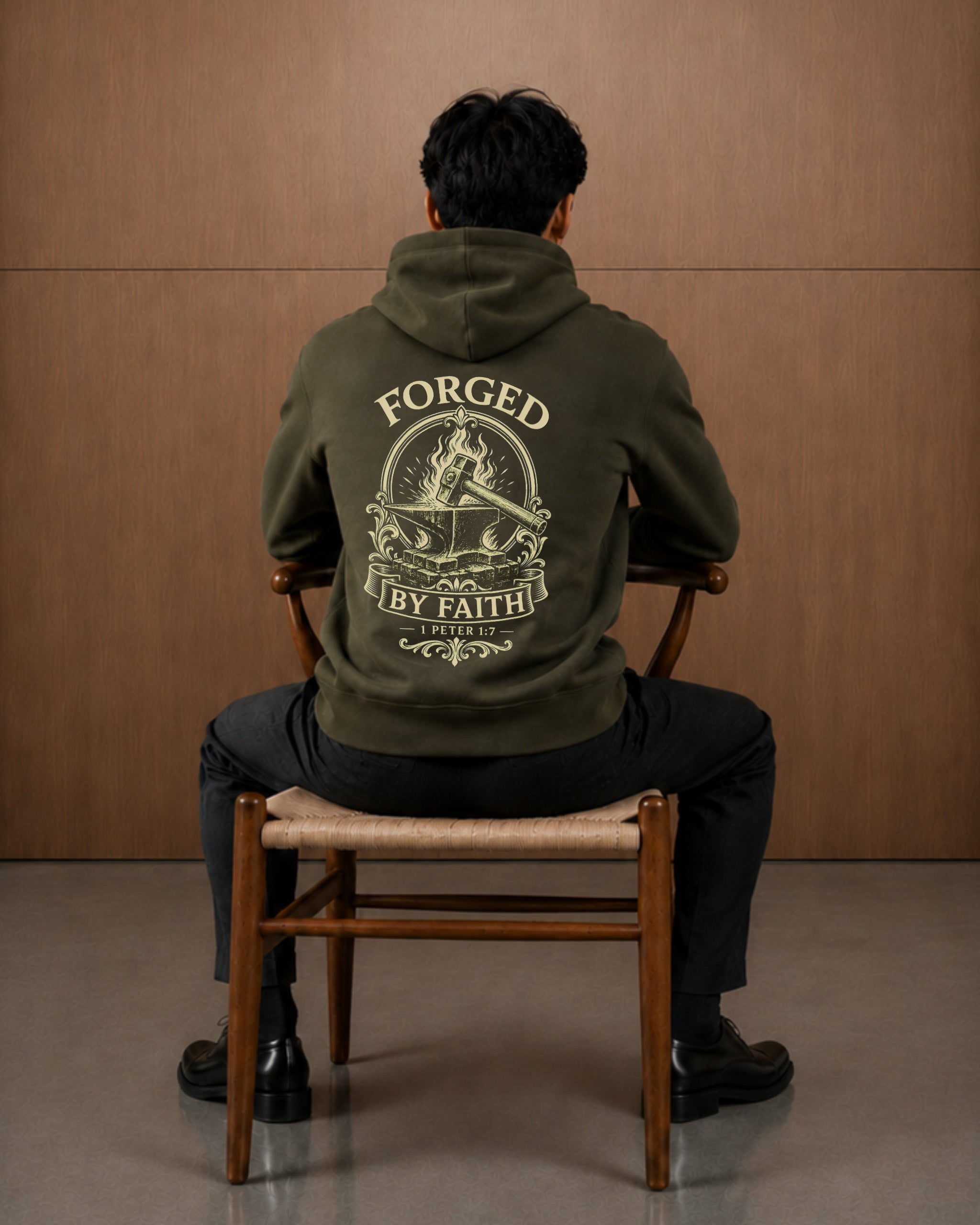 A man sits on a wooden chair facing away from the camera, wearing an olive green hoodie with a large cream graphic on the back that reads 'FORGED BY FAITH'.