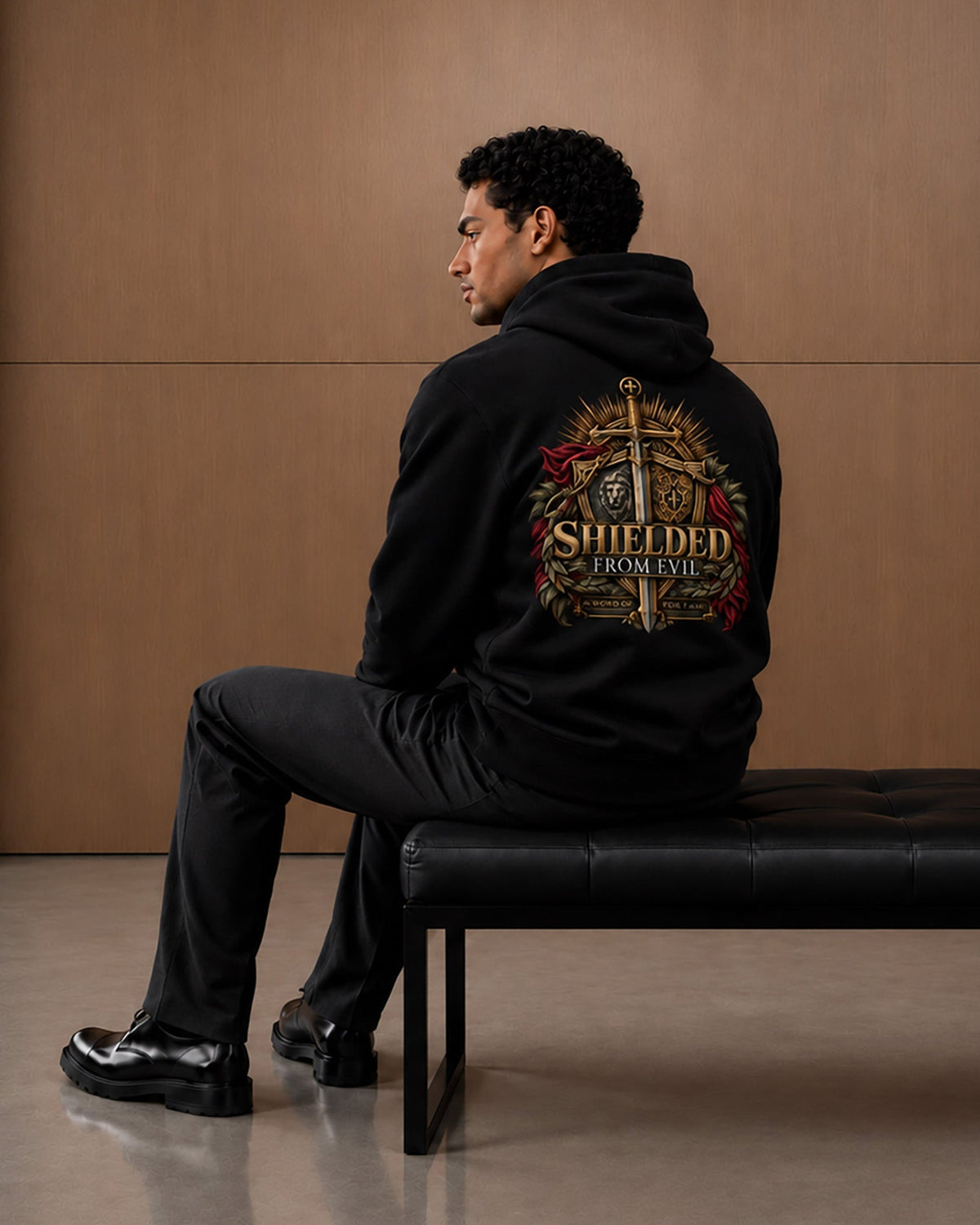 A man wearing a black hoodie with a large 'The Shielded From Hell' graphic on the back, sitting on a black leather bench against a wood-paneled wall.
