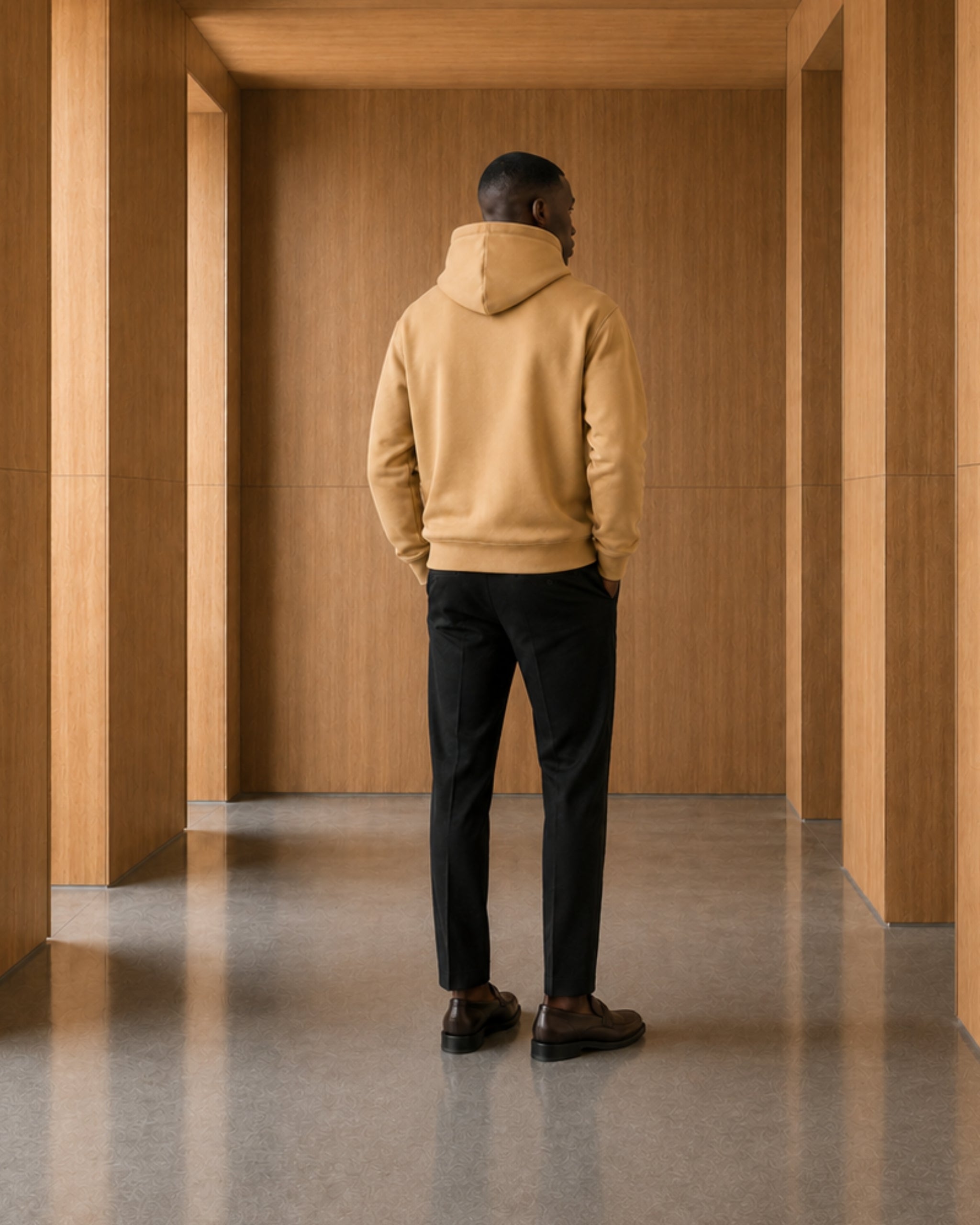 Man wearing a beige hoodie and black pants standing in a wooden architectural space.
