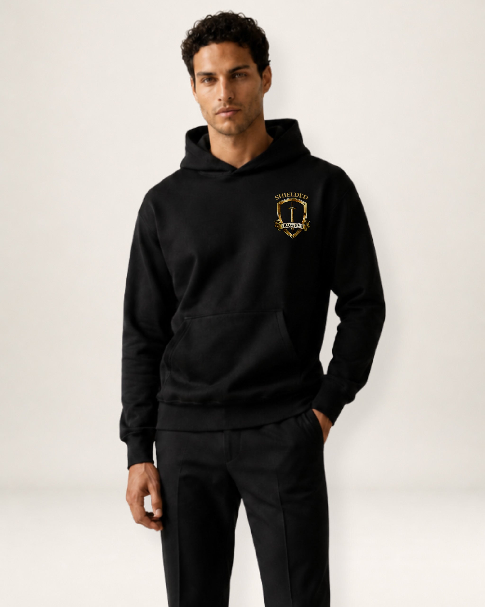 A man wearing a black pullover hoodie with a gold and black crest graphic on the left chest, paired with black sweatpants, photographed against a clean off-white studio background.