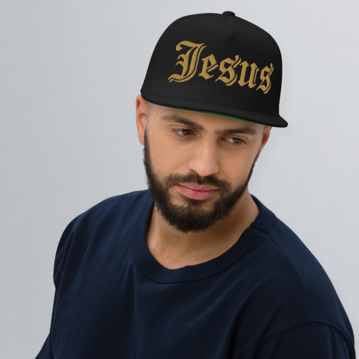 A man wearing a black snapback cap with 'Jesus' embroidered in a gold Gothic font, paired with a dark navy t-shirt, photographed against a neutral gray background.