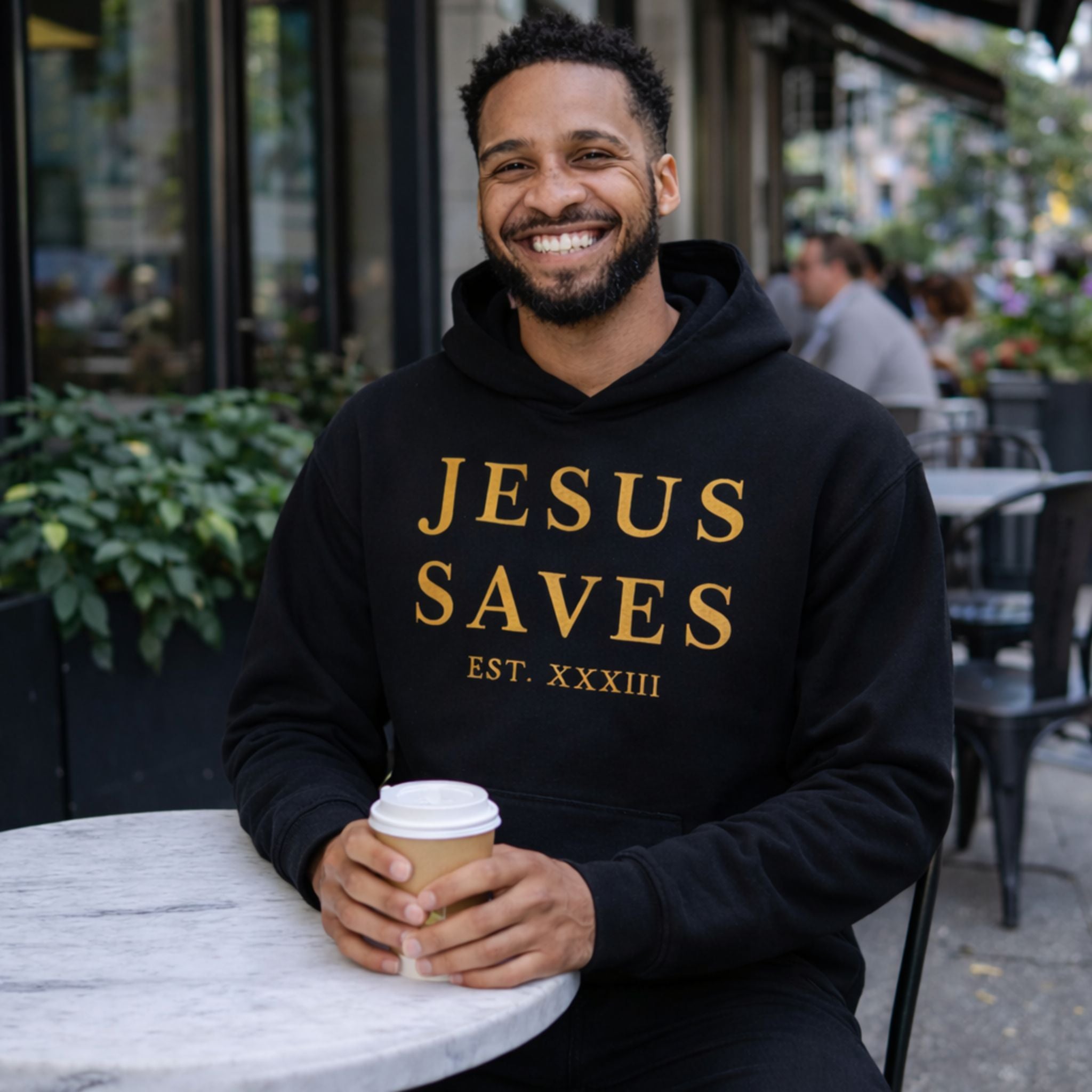 Man wearing a black hoodie with 'JESUS SAVES' text, holding a coffee cup outdoors.