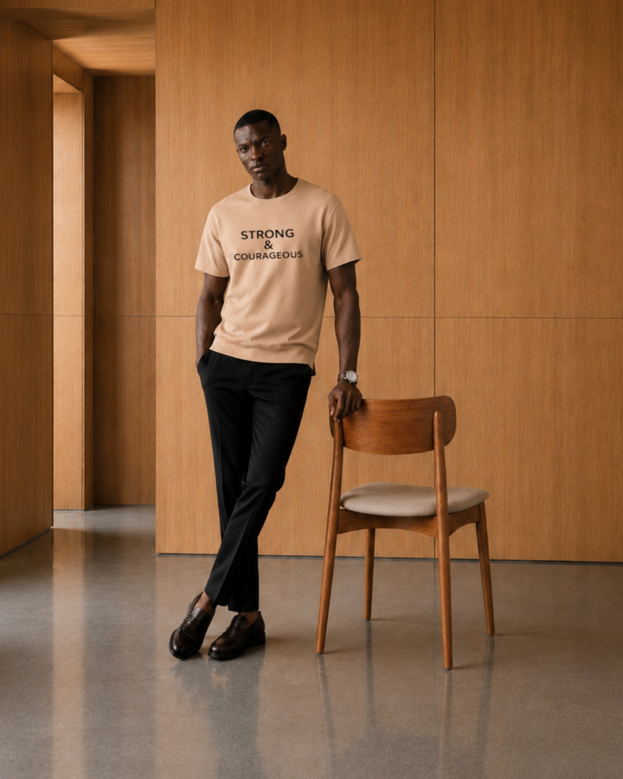 Man wearing a beige t-shirt with text standing next to a wooden chair in a minimalistic room.